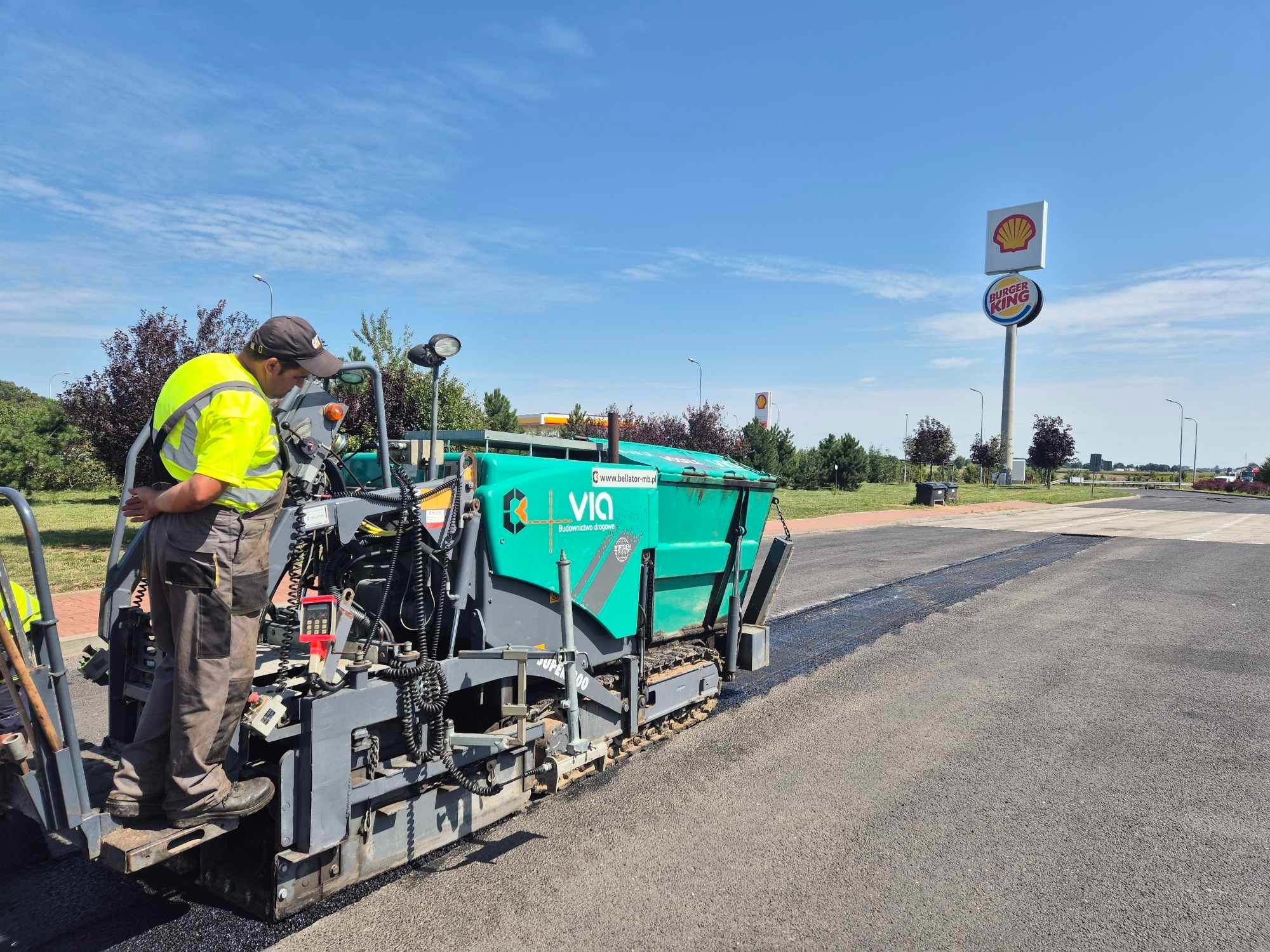Pracownik w odblaskowej kamizelce obsługuje rozściełacz asfaltu VIA na budowie drogi, w tle widoczny znak Shell i Burger King.