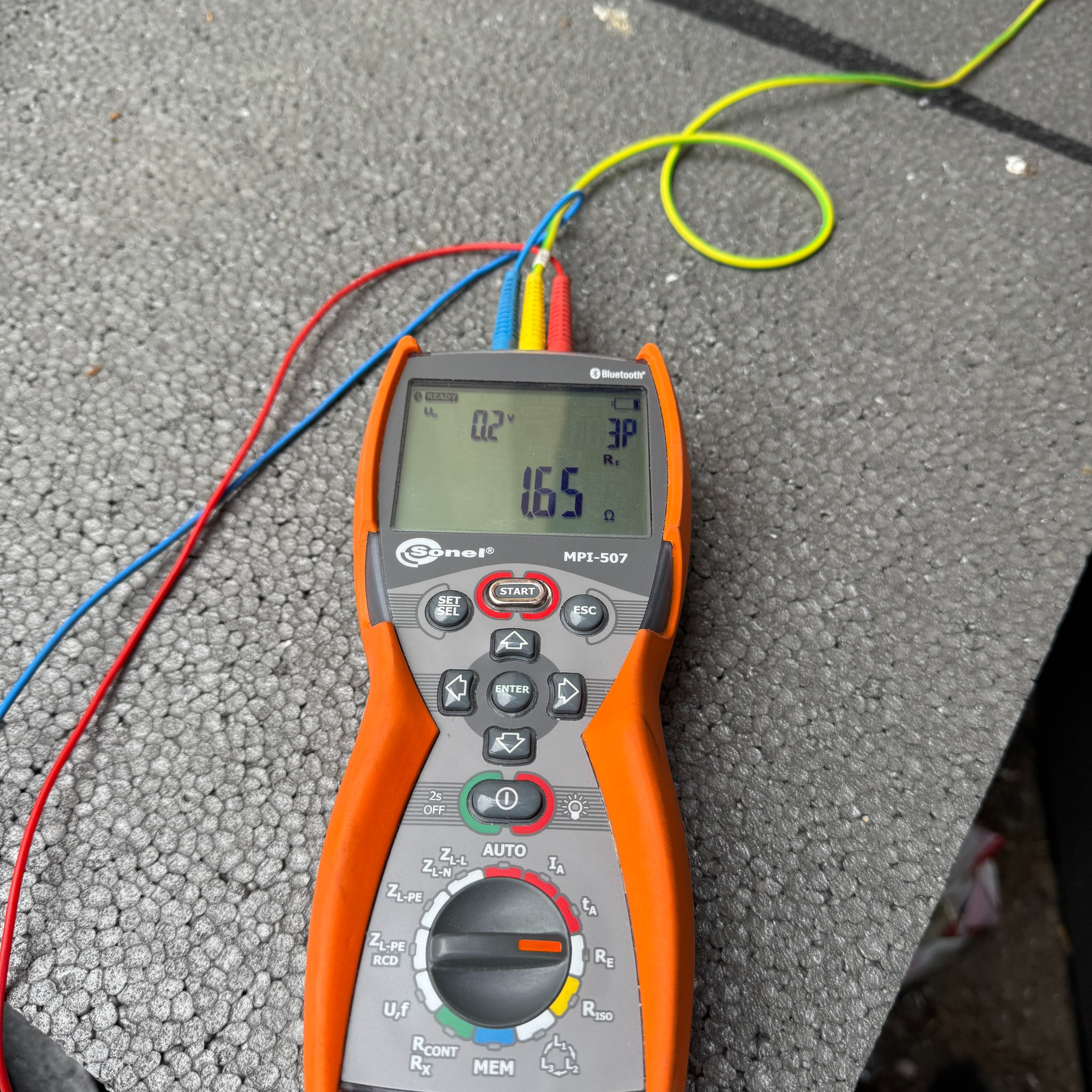 Pomarańczowy miernik Sonel MPI-507 z podłączonymi przewodami pomiarowymi na szorstkiej powierzchni. Wyświetlacz pokazuje wartości pomiarowe.