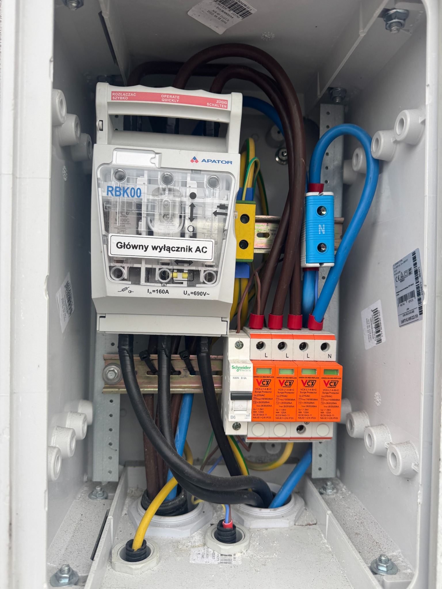 Rozdzielnica elektryczna z bezpiecznikami i wyłącznikiem AC w białej obudowie. Widoczne przewody w różnych kolorach i oznaczenia. Instalacja fotowoltaiczna.