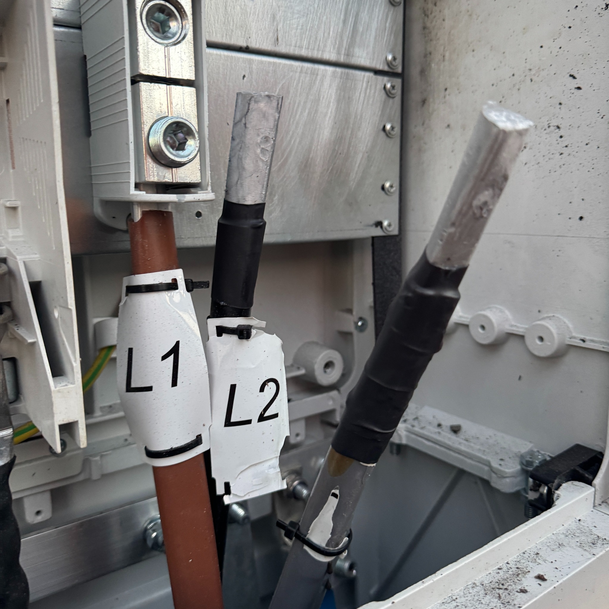 Instalacja elektryczna w skrzynce: kable L1, L2 i neutralny, złącza śrubowe, widoczne przewody uziemiające. Detal montażu instalacji fotowoltaicznej.