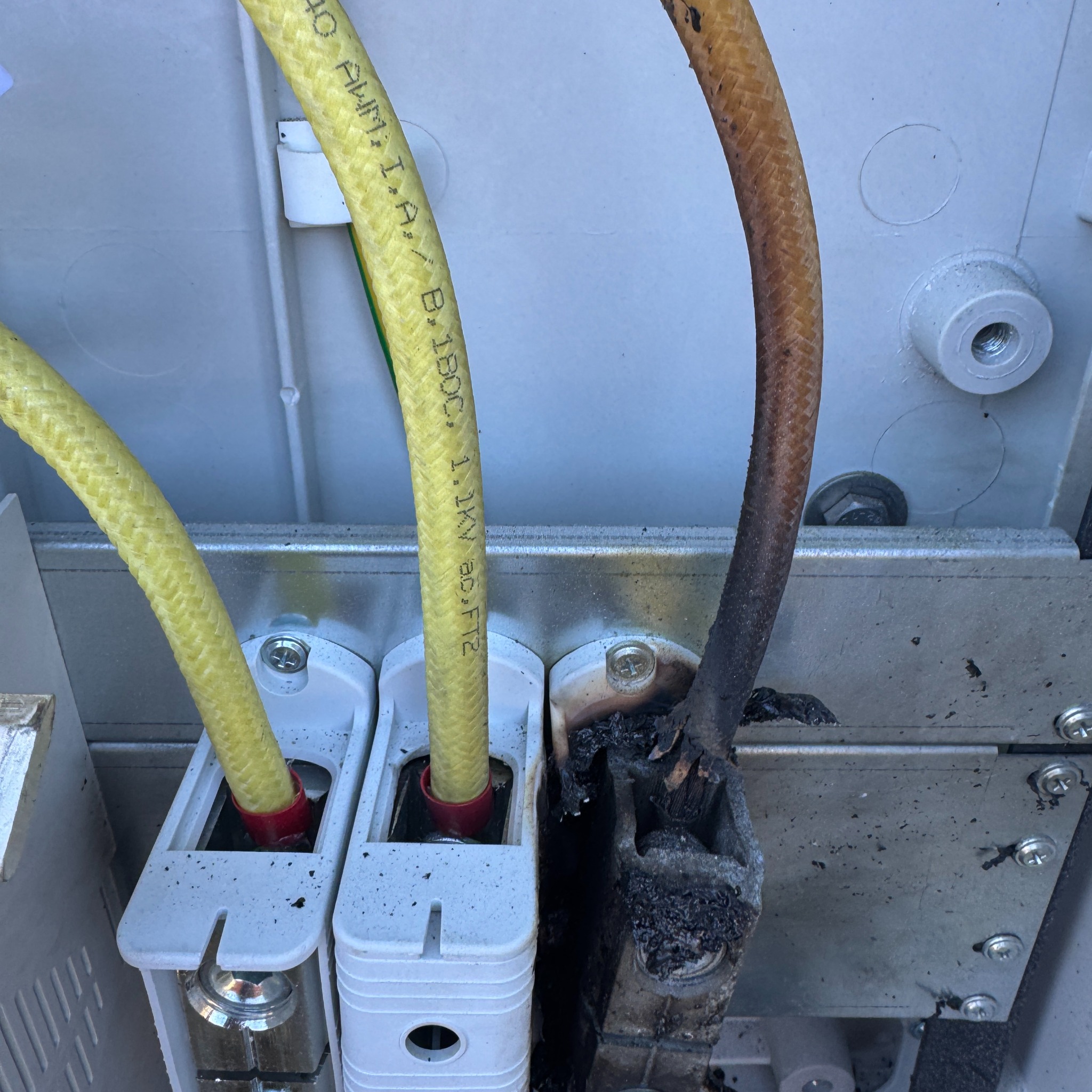 Spalona instalacja fotowoltaiczna w skrzynce przyłączeniowej. Uszkodzony przewód, zwęglona obudowa. Żółte przewody z oznaczeniami. Niebezpieczny stan instalacji.