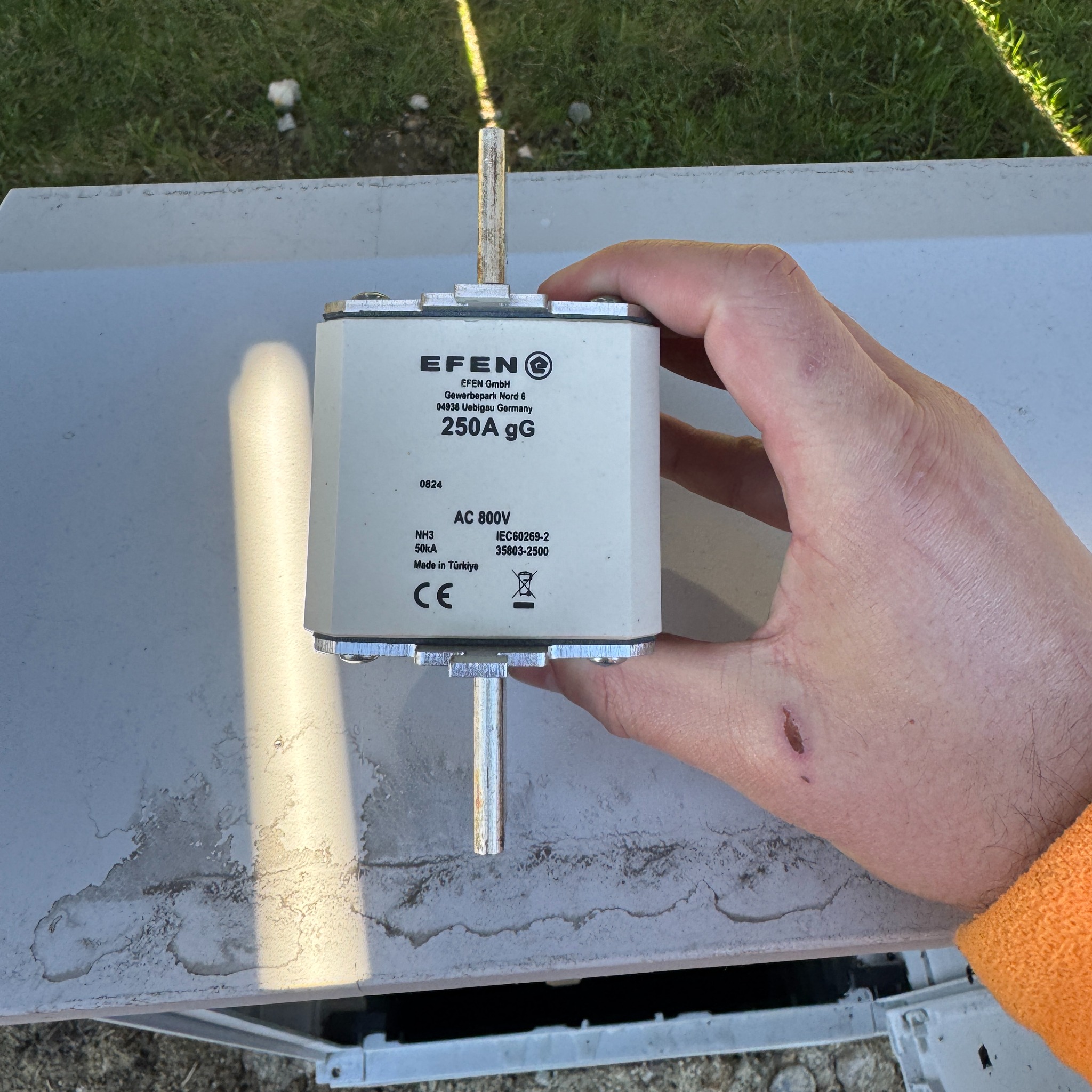 Ręka trzyma bezpiecznik EFEN 250A gG AC 800V nad szarą skrzynką. Widoczne oznaczenia i certyfikaty na bezpieczniku. Element instalacji fotowoltaicznej.