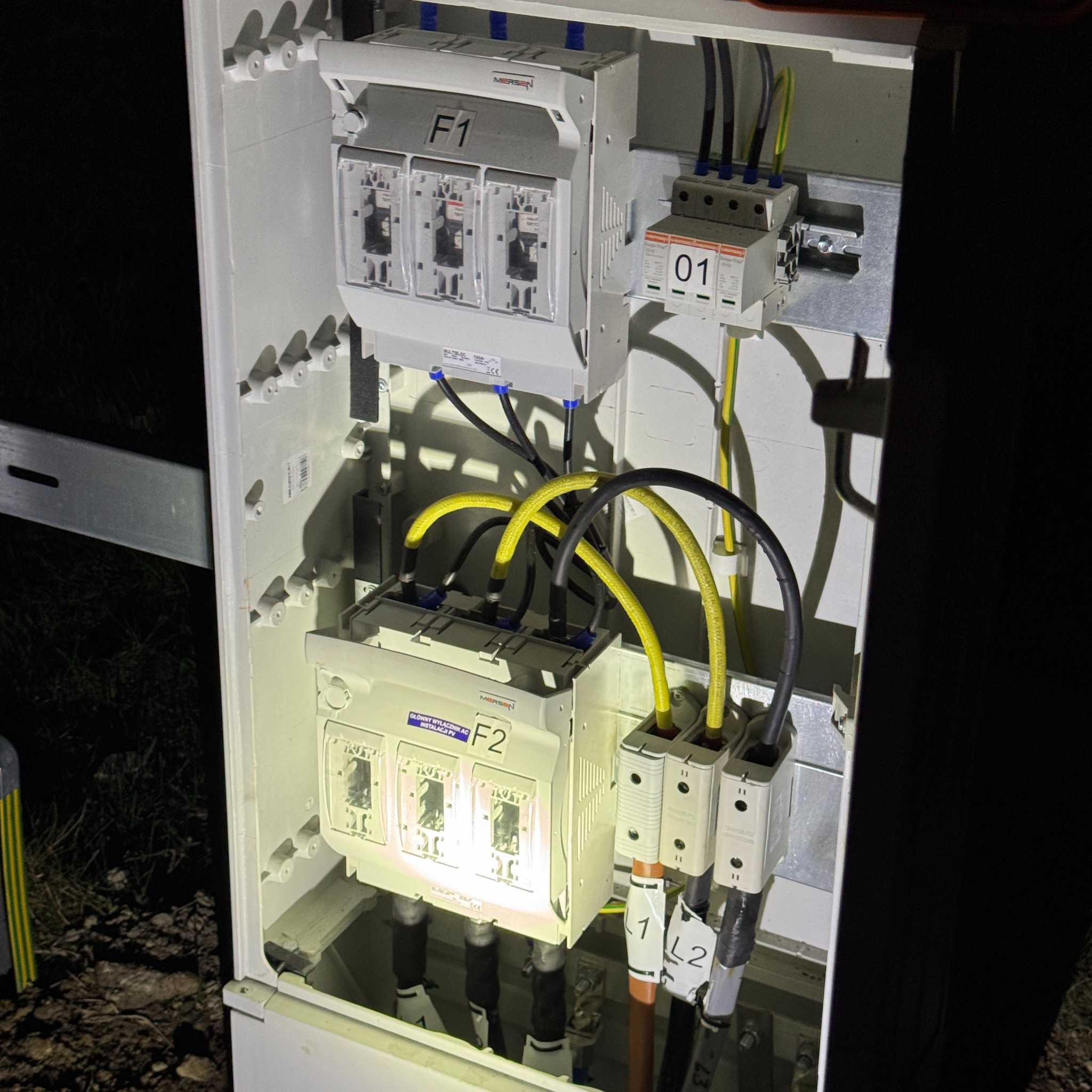 Wnętrze skrzynki elektrycznej z bezpiecznikami, wyłącznikami i okablowaniem instalacji fotowoltaicznej, widoczne oznaczenia F1 i F2 oraz przewody w kolorach żółtym i czarnym.