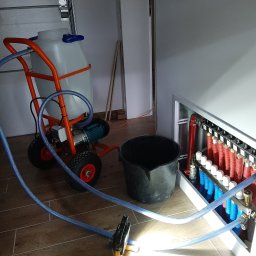 Oleksandr Borodavkin - Instalacja rozdzielacza ogrzewania podłogowego z użyciem wózka z pompą i zbiornikiem, widoczne podłączenia rur w kolorach czerwonym i niebieskim, oraz węże doprowadzające wodę.