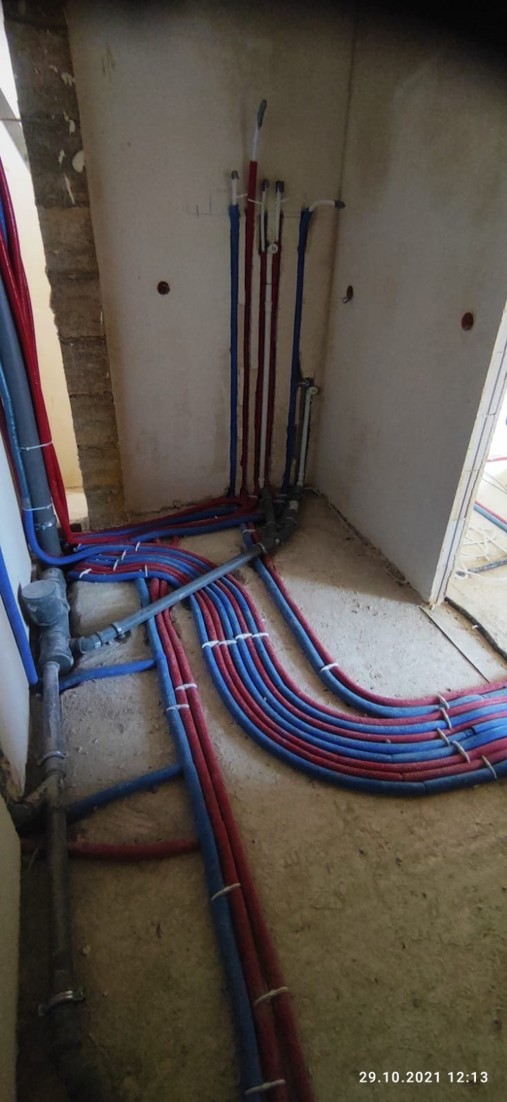 Instalacja hydrauliczna w surowym stanie: ułożone rury czerwone i niebieskie na podłodze, pionowe rury przy ścianie, widoczne łączenia i mocowania, surowa ściana w tle.