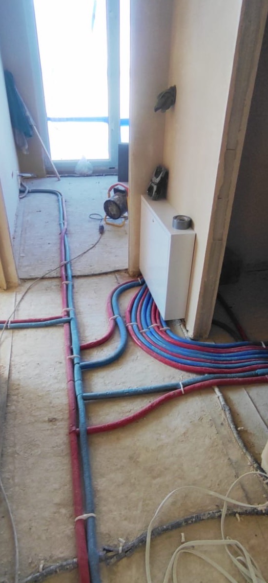 Instalacja ogrzewania podłogowego w trakcie montażu z widocznymi rurami w kolorach czerwonym i niebieskim, prowadzonymi po szlichtcie, przymocowanymi opaskami, z rozdzielaczem na ścianie...