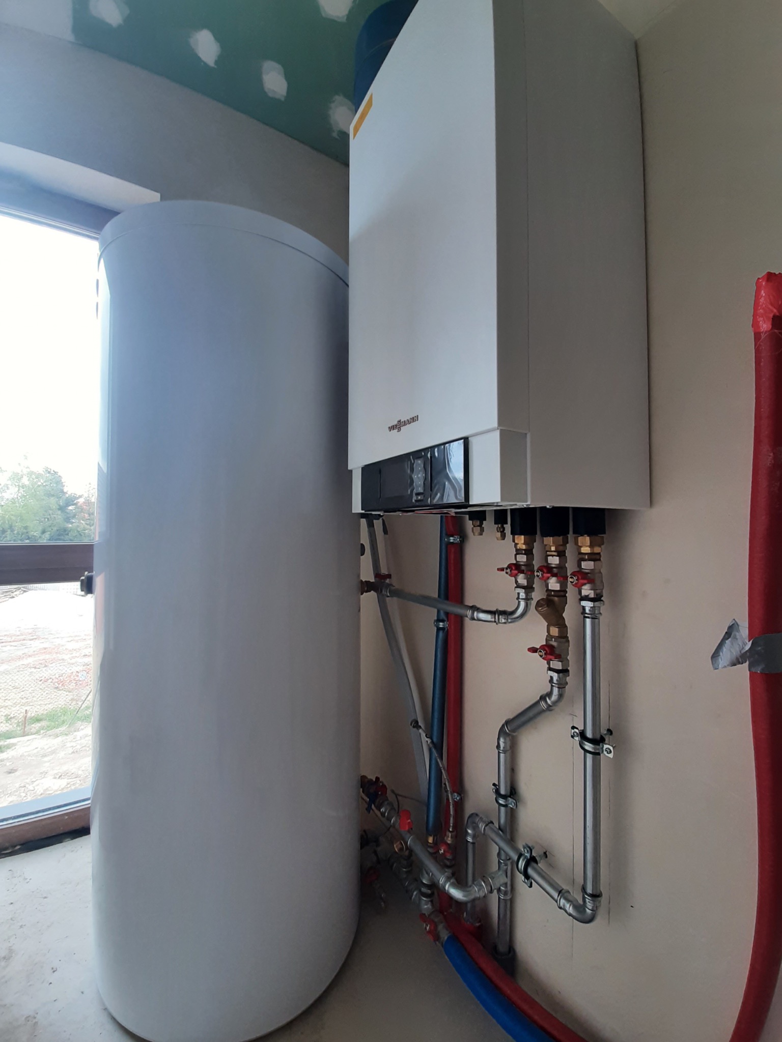 Biały piec CO wiszący na ścianie z podłączonymi rurami miedzianymi i zbiornikiem na wodę, widoczne okno w tle, ściany w stanie surowym.