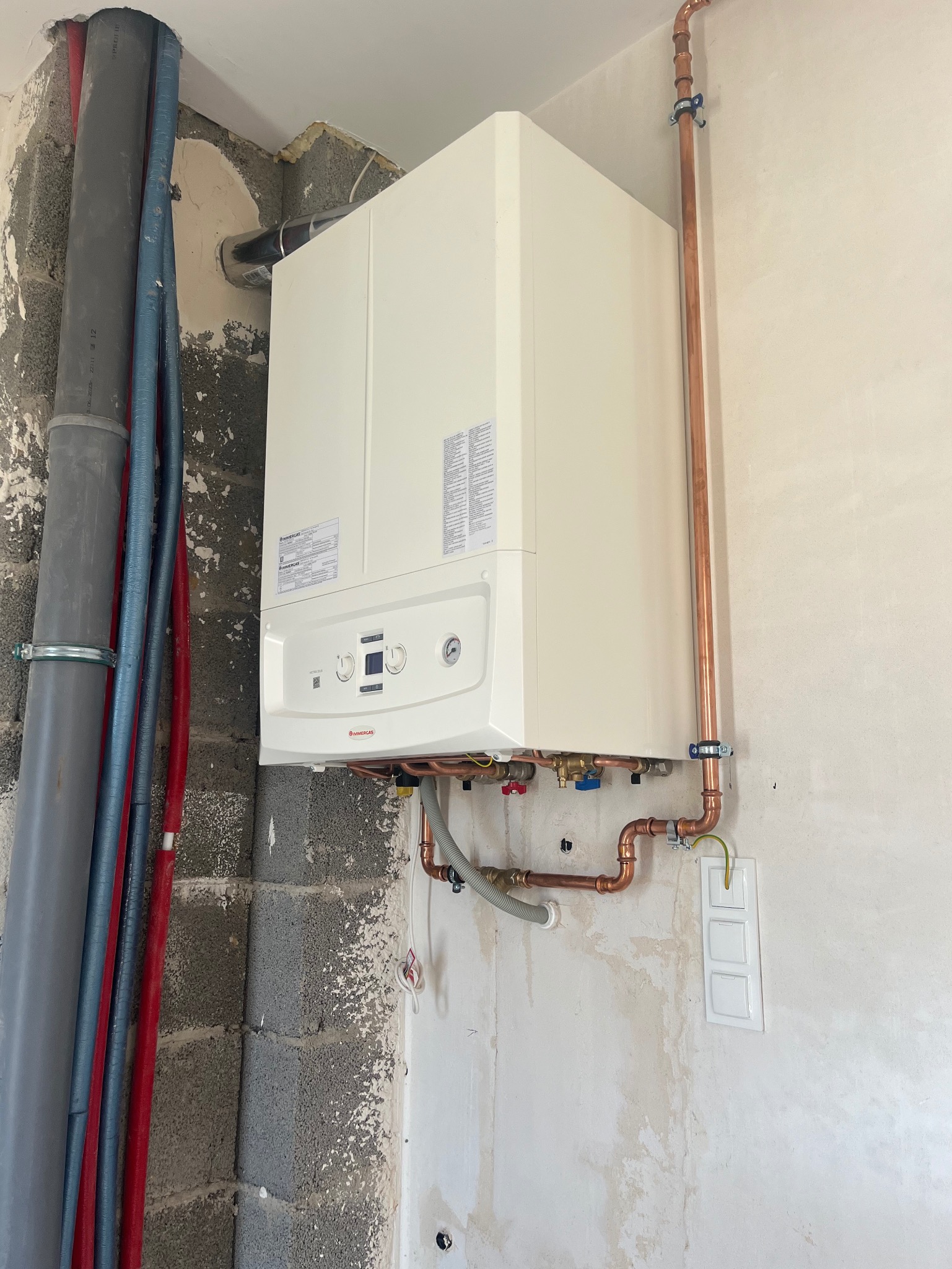 Nowy, biały piec centralnego ogrzewania zamontowany na ścianie w surowym stanie, z widocznymi miedzianymi rurami i szarymi rurami plastikowymi, przed wykończeniem ściany.