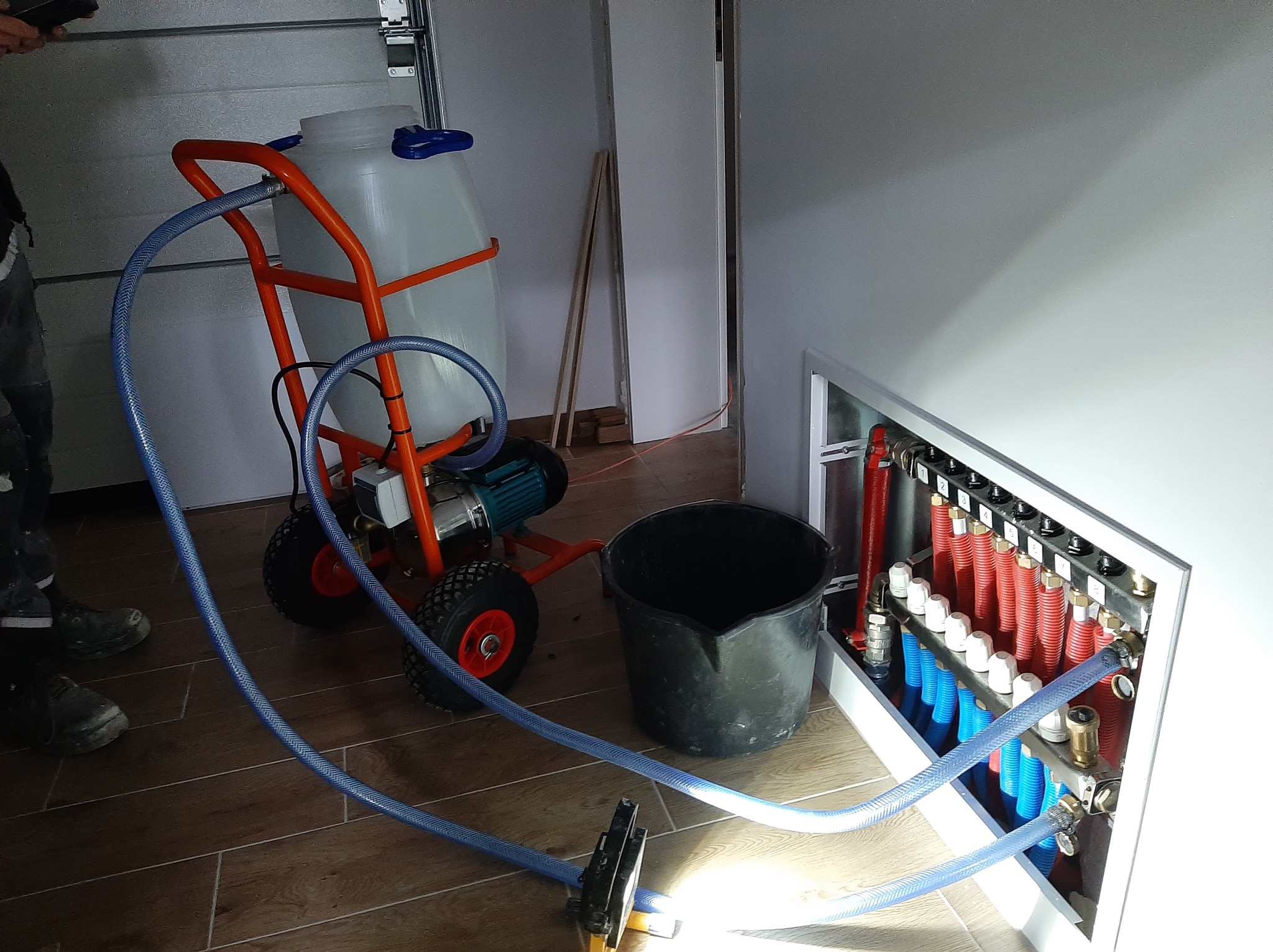 Instalacja rozdzielacza ogrzewania podłogowego z użyciem wózka z pompą i zbiornikiem, widoczne podłączenia rur w kolorach czerwonym i niebieskim, oraz węże doprowadzające wodę.