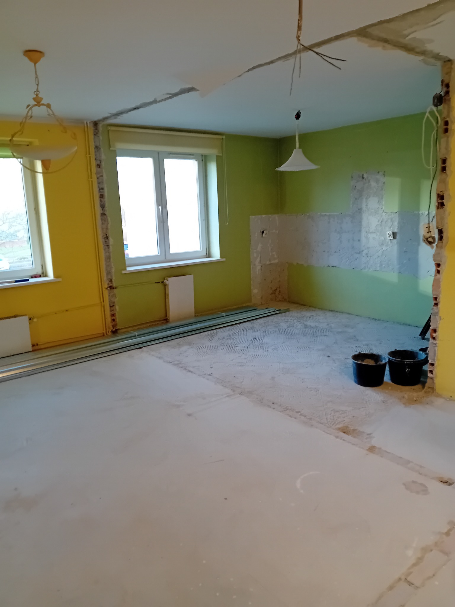 Remont kuchni w Bogatyni: przygotowanie do położenia płytek, widoczne ślady po starych płytkach, dwa wiadra i listwy przypodłogowe. Żółto-zielone ściany.