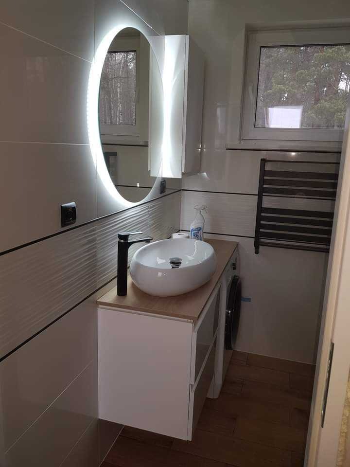 Nowoczesna łazienka z owalną umywalką nablatową, podświetlanym lustrem i drewnianą podłogą. Widoczna pralka i grzejnik drabinkowy.