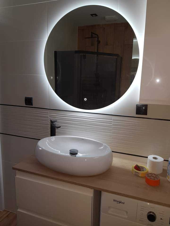 Nowoczesna łazienka z okrągłym lustrem LED, owalną umywalką nablatową i pralką pod blatem. Białe płytki z subtelną fakturą i czarna armatura.
