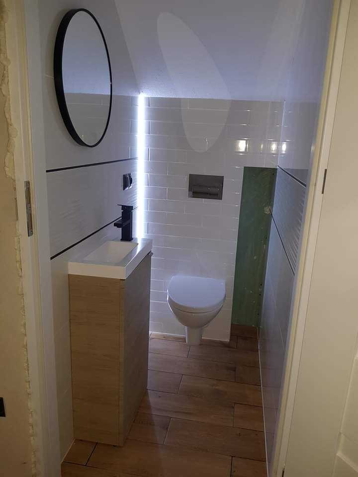 Wykończona, wąska łazienka z umywalką na szafce, toaletą i okrągłym lustrem. Białe płytki na ścianach, drewnopodobne panele na podłodze. Oświetlenie LED w pionowej szczelinie.