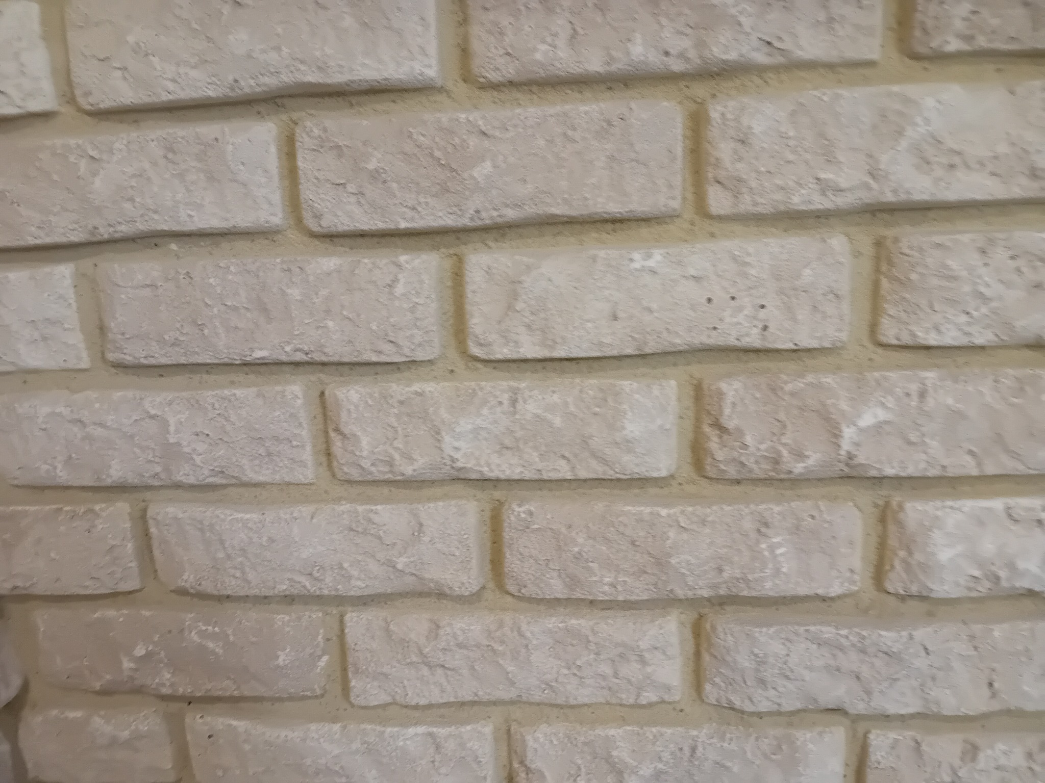 Ściana wykończona dekoracyjną, beżową cegłą z wyraźną fakturą. Widoczne spoiny między cegłami. Ujęcie z bliska, ukazujące detale materiału.