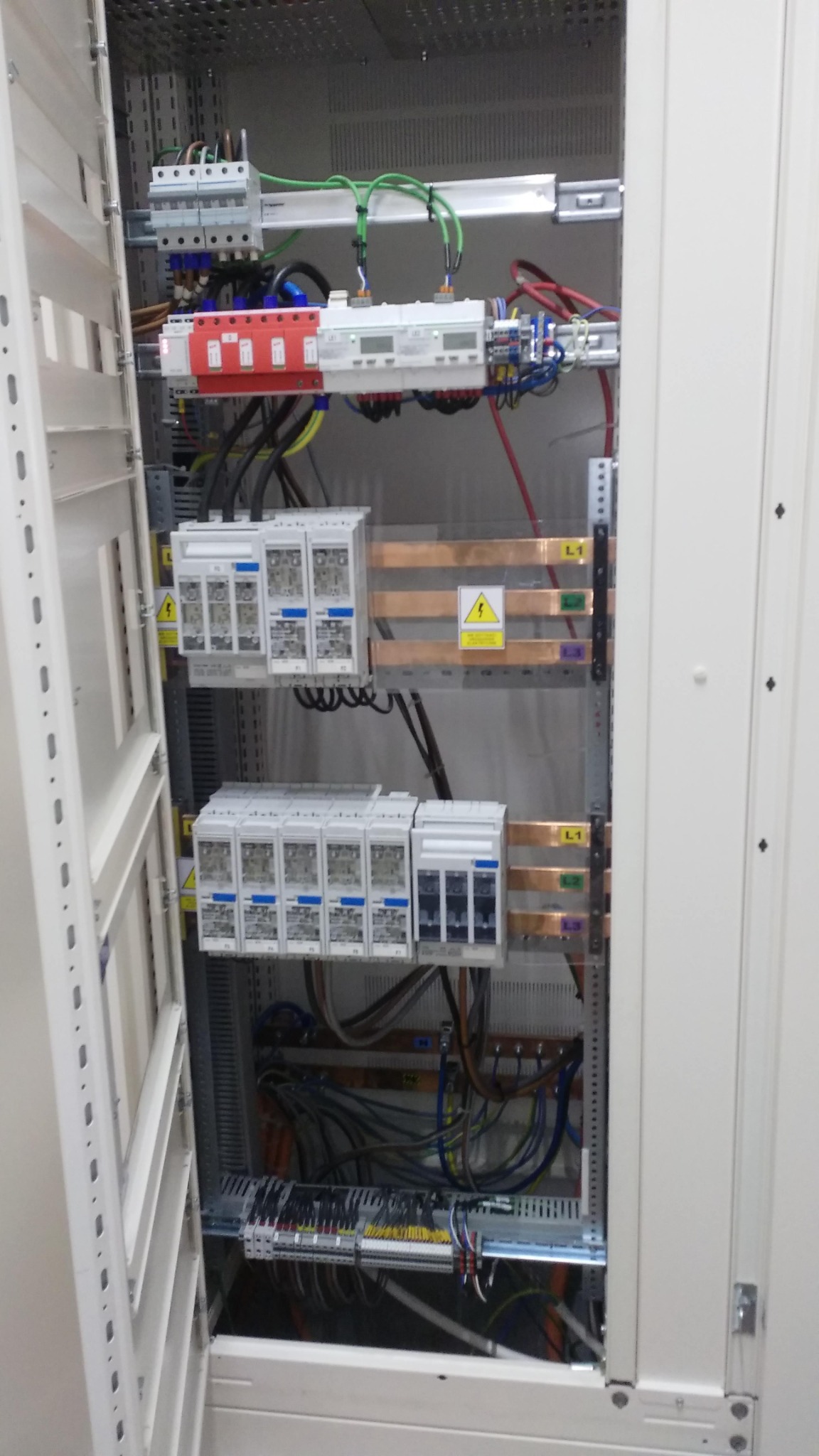 Otwarta szafa elektryczna z widocznymi zabezpieczeniami, przewodami i szynami miedzianymi, etykiety ostrzegawcze o napięciu, widok od frontu na wnętrze rozdzielnicy.