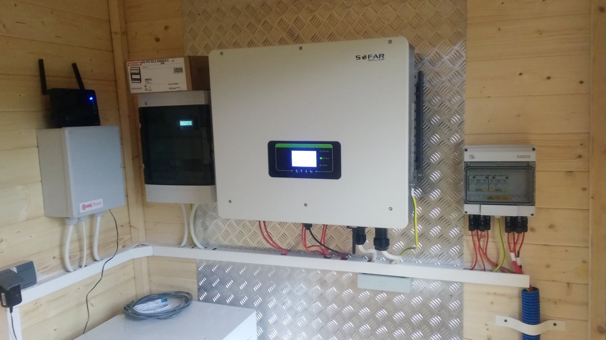 Zamknięta przestrzeń z panelami elektrycznymi, inwerterem Sofar Solar z wyświetlaczem, routerem i przewodami na ścianach z drewnianych desek i blachy ryflowanej.