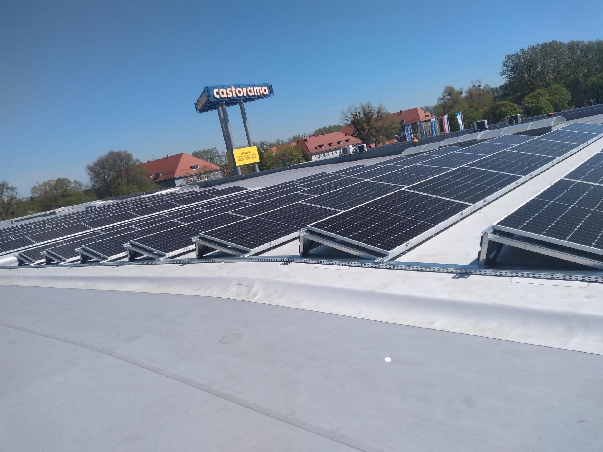 Rząd paneli słonecznych na szarym dachu płaskim z widocznym znakiem Castorama w tle, w słoneczny dzień.