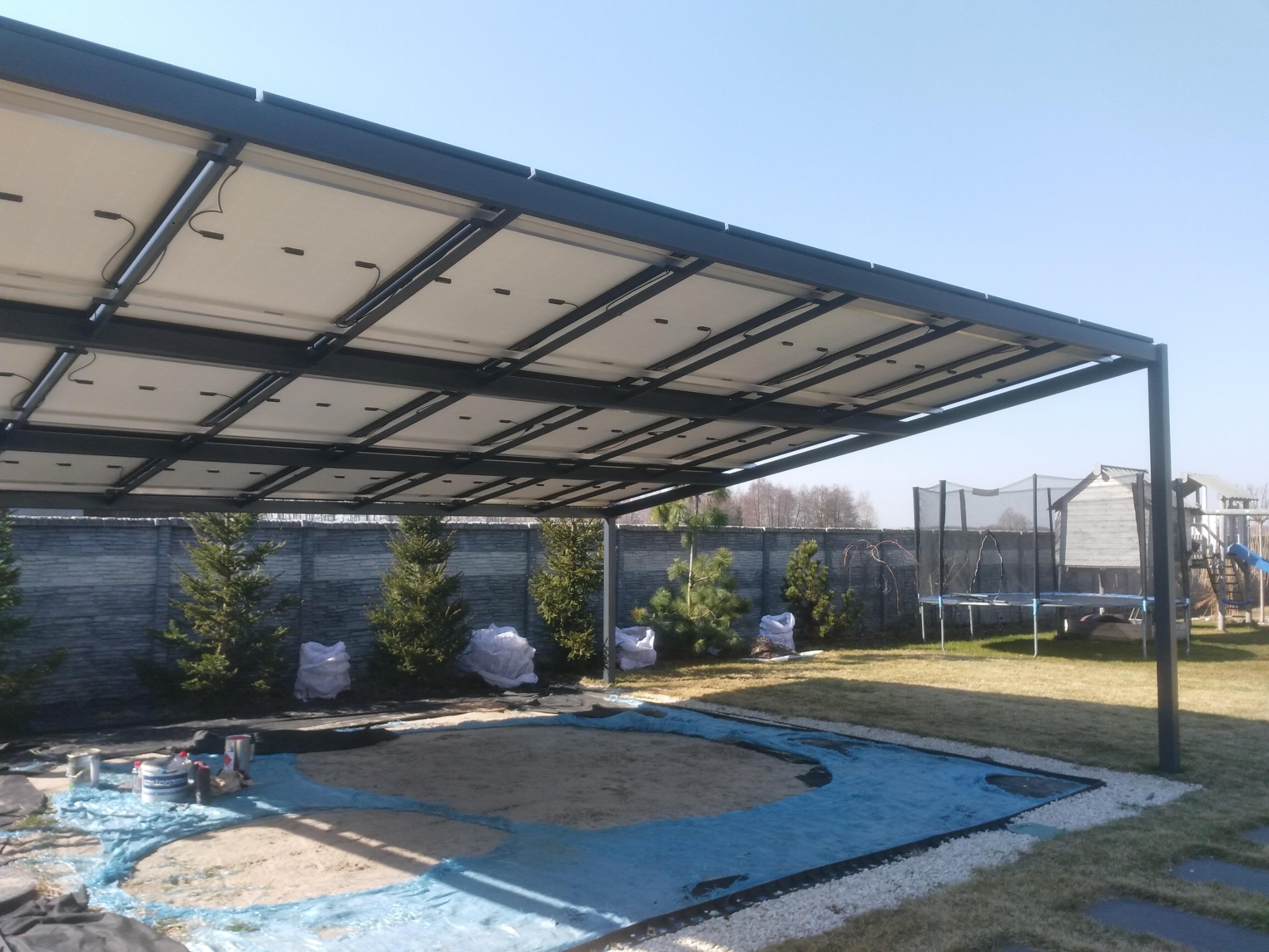 Pergola z panelami fotowoltaicznymi nad przygotowanym wykopem, zabezpieczonym folią, z widocznymi puszkami farb i workami z ziemią w tle, na trawniku.