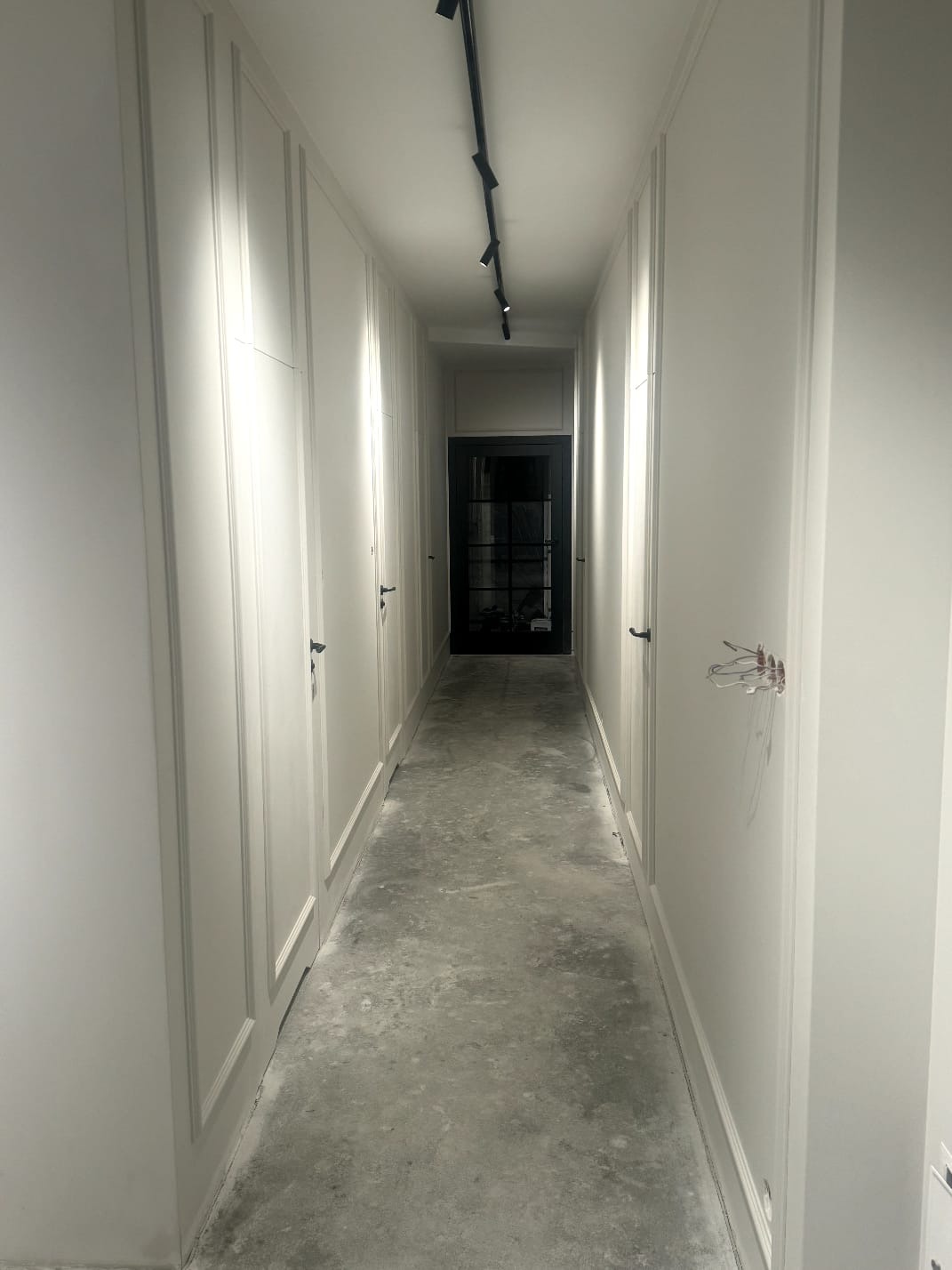 Długi korytarz z białymi szafami w zabudowie po obu stronach, surowa podłoga, czarne oświetlenie szynowe na suficie i czarne drzwi na końcu korytarza, widoczne przewody elektryczne.