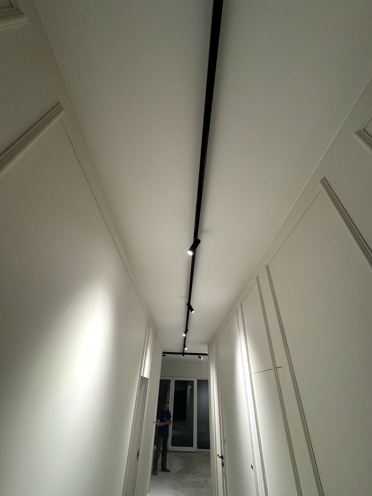Długi korytarz z białymi ścianami z dekoracyjnymi listwami i czarnym szynoprzewodem z reflektorami punktowymi, widoczna sylwetka osoby w tle.