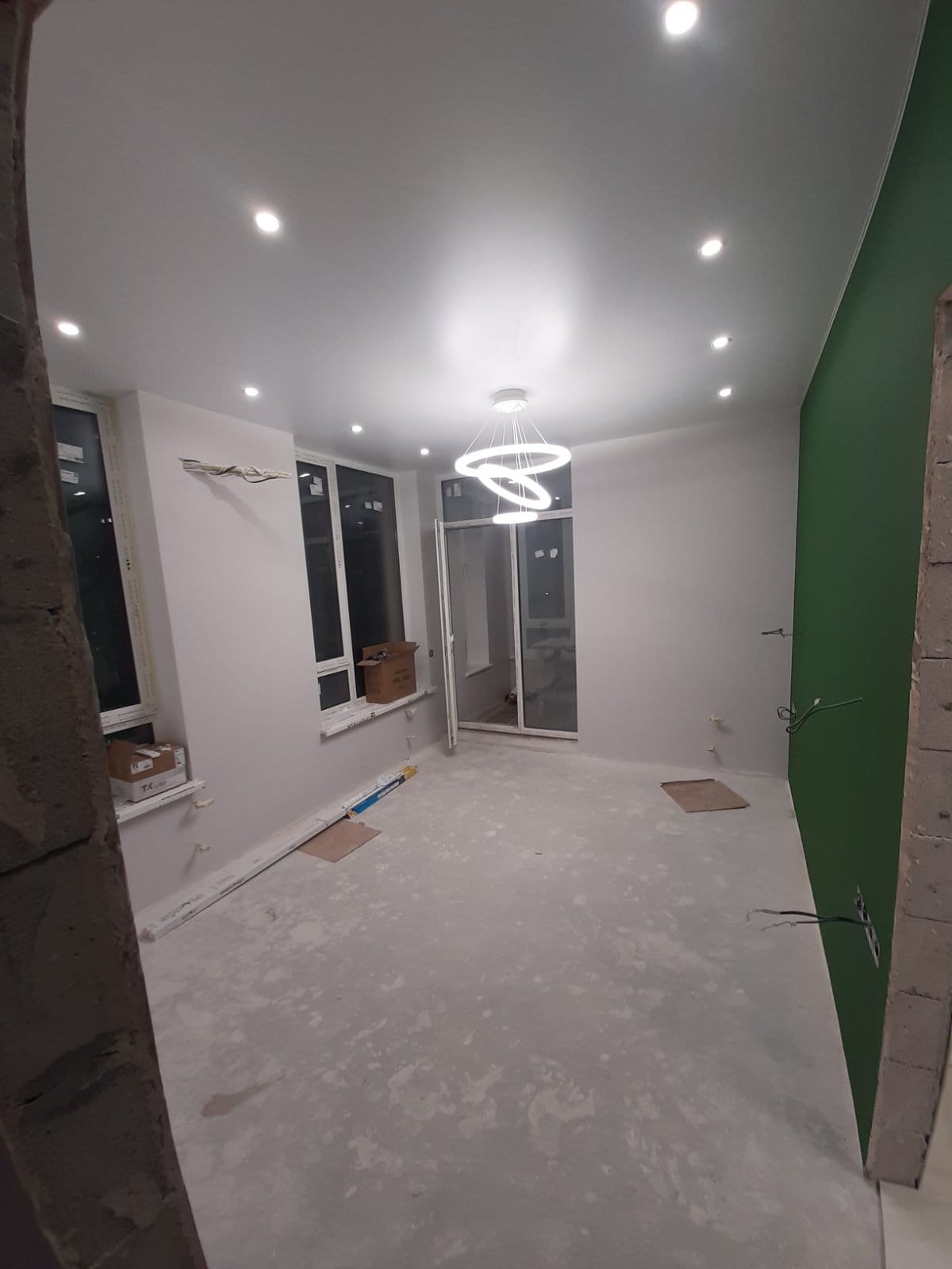 Pokój w trakcie prac wykończeniowych: świeżo pomalowane ściany w odcieniach szarości i zieleni, nowoczesna lampa wisząca z trzema okręgami, widoczne puszki elektryczne i przewody, podłoga...