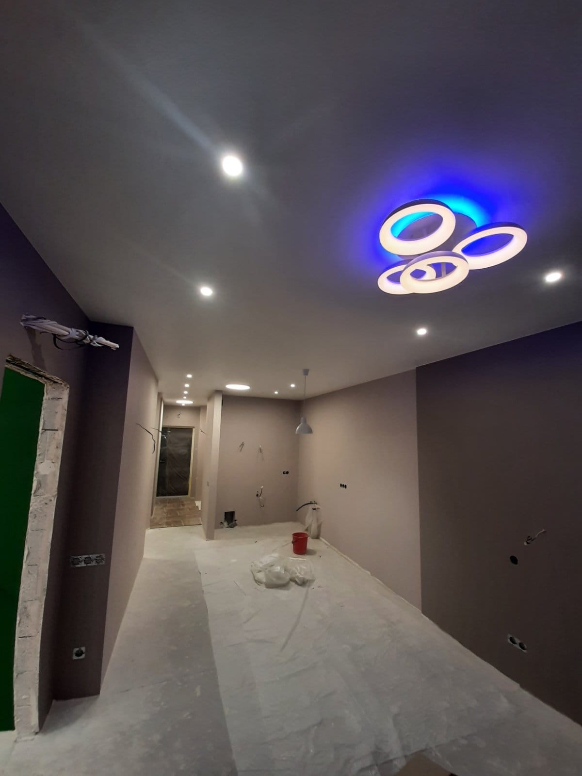 Wnętrze w trakcie prac wykończeniowych: ściany pomalowane na szaro, podłoga zabezpieczona folią, nowoczesna lampa sufitowa z niebieskim podświetleniem, widoczne punkty elektryczne.