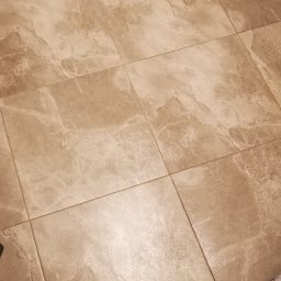 Marian Jankowski - Beżowe, matowe płytki podłogowe w łazience, ułożone w kwadratowy wzór, z widocznymi fugami i fragmentem szarej tkaniny w prawym dolnym rogu.