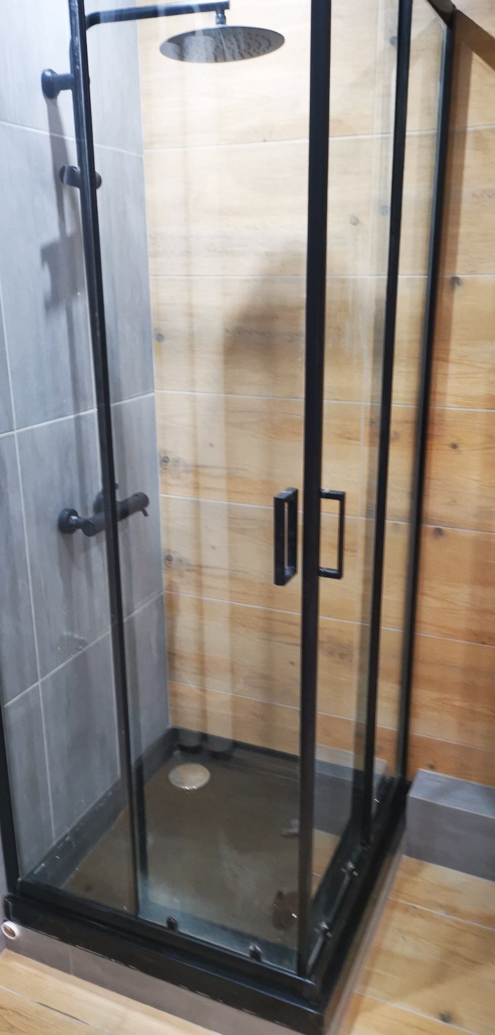 Nowoczesna kabina prysznicowa z czarnymi profilami, szklane ściany, deszczownica, szare płytki i drewnopodobne panele na ścianach.