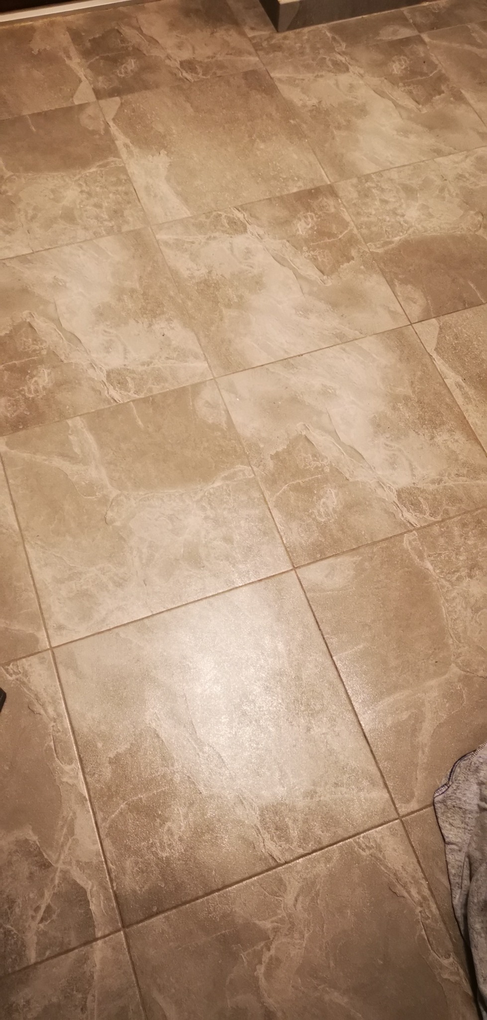 Beżowe, matowe płytki podłogowe w łazience, ułożone w kwadratowy wzór, z widocznymi fugami i fragmentem szarej tkaniny w prawym dolnym rogu.