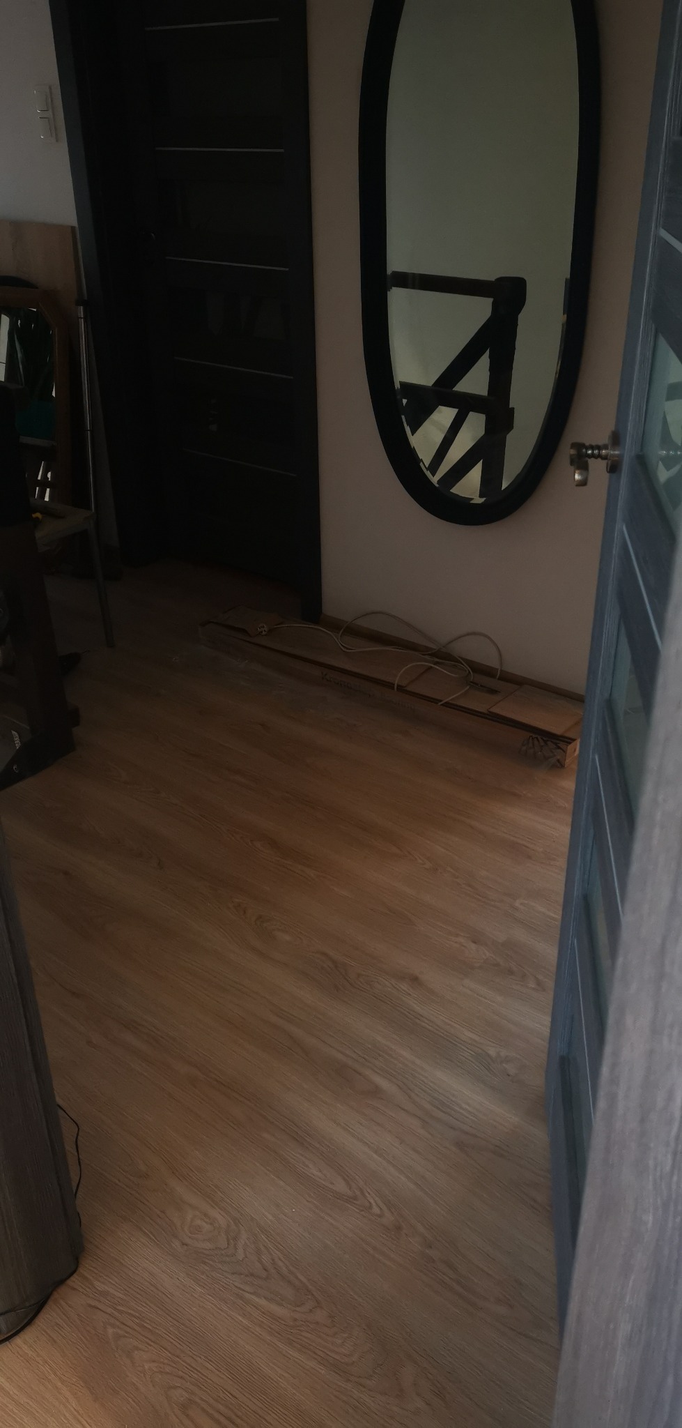 Nowo ułożone panele podłogowe w odcieniach brązu, widoczne kable elektryczne i listwa przypodłogowa przed montażem, czarne drzwi w tle i odbicie schodów w owalnym lustrze.