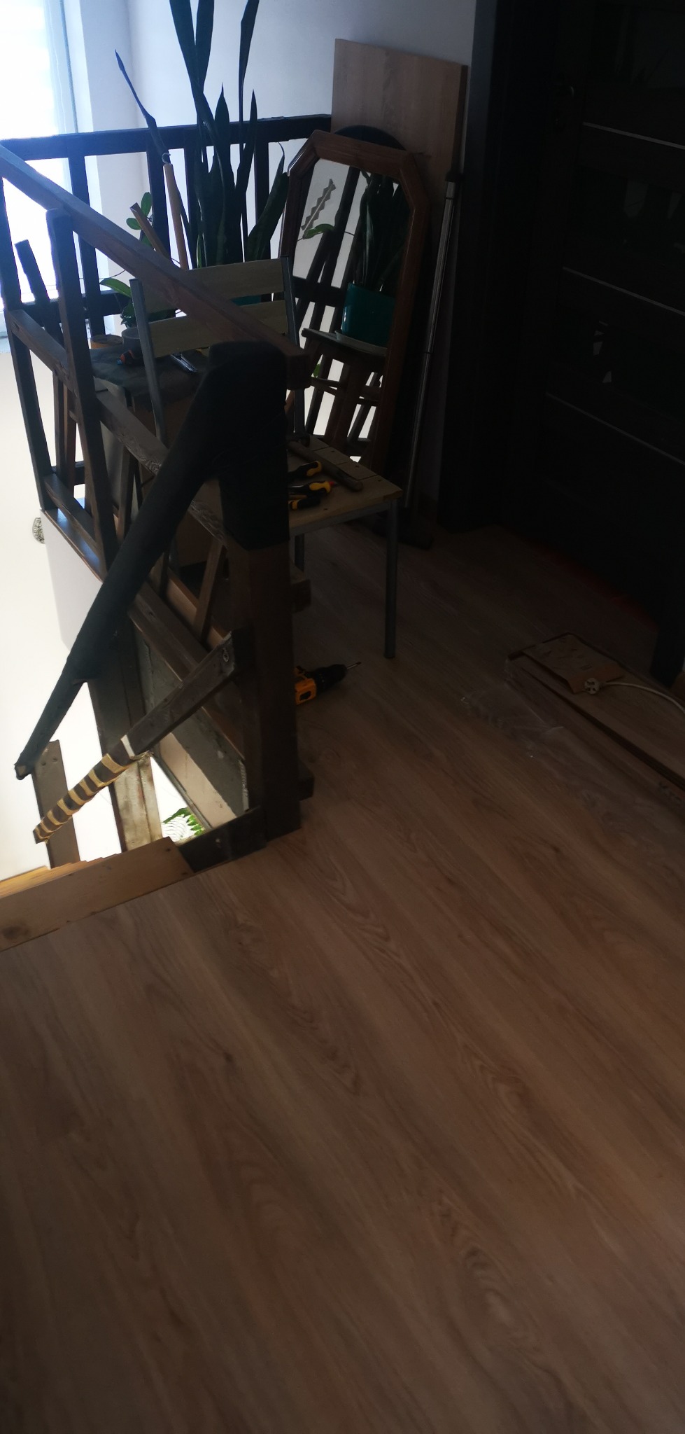 Fragment drewnianej balustrady schodów w trakcie montażu, widoczna wiertarka i ułożone panele podłogowe, w tle rośliny doniczkowe i lustro.