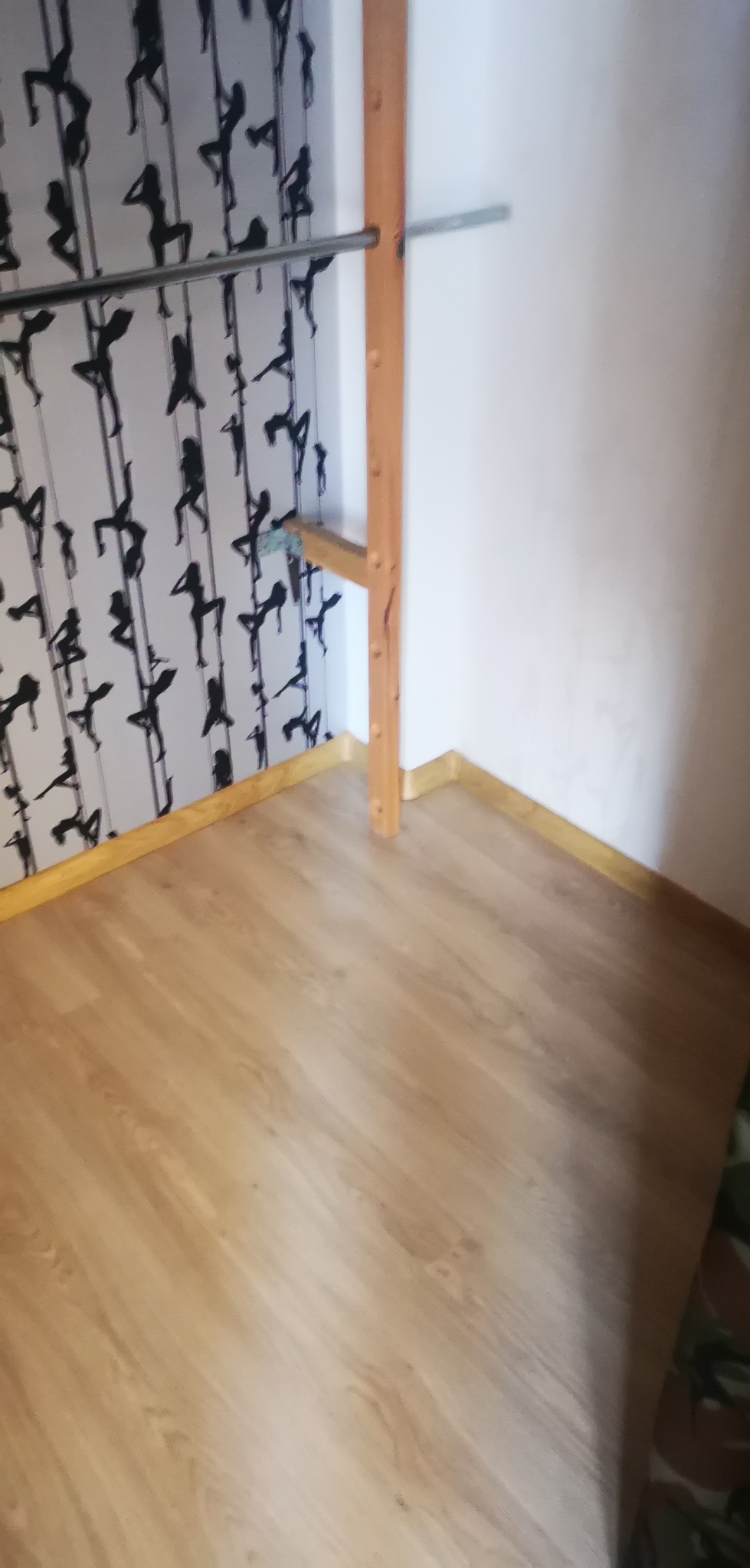 Fragment pokoju po remoncie: jasne panele podłogowe, ściana z tapetą w czarne sylwetki tancerek na rurze, drewniany element konstrukcyjny przy ścianie.