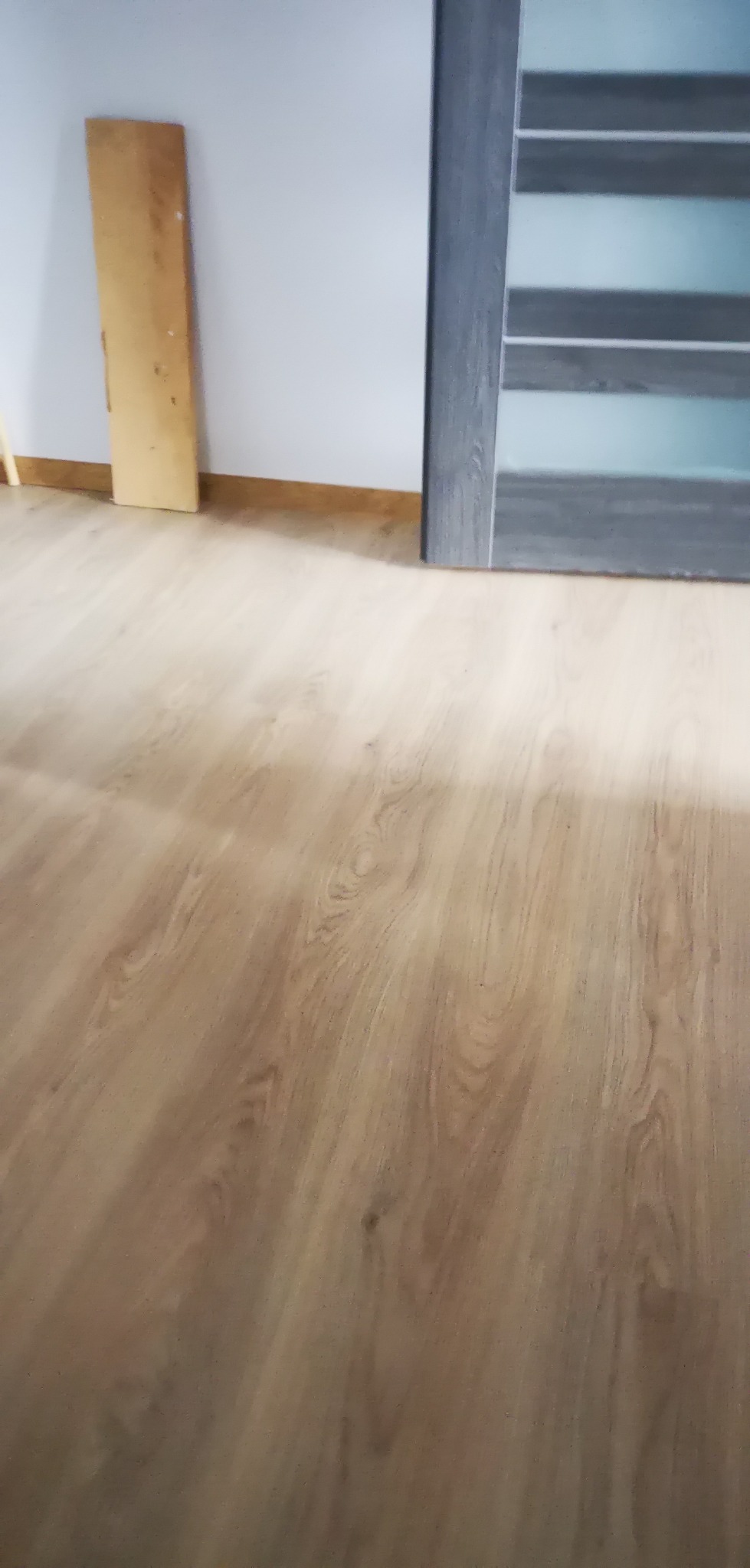 Jasne panele podłogowe z widoczną fakturą drewna, fragment drzwi z szarą ościeżnicą i szybami, oraz pionowy element z surowego drewna przy białej ścianie.