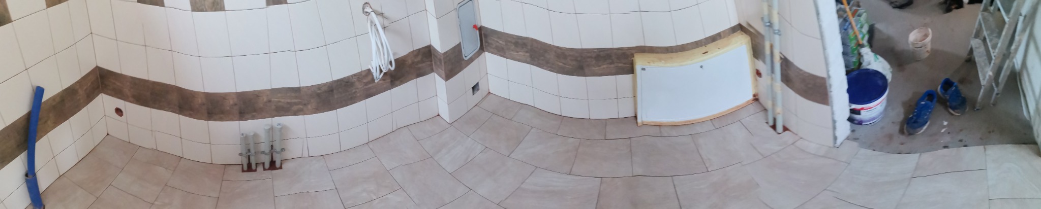 Panoramiczne ujęcie wnętrza w trakcie prac wykończeniowych, widoczne ułożone płytki podłogowe i ścienne z dekoracyjnym pasem, wystające rury instalacji, przygotowane miejsce na otwór rewizyjny...