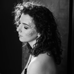 STUDIO KREATYWNE Katarzyna Ćwiertnia - Czarno-białe portretowe ujęcie profilu kobiety z kręconymi włosami i brokatem na twarzy, w zamyślonej pozie na tle ciemnych zasłon.