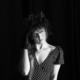 STUDIO KREATYWNE Katarzyna Ćwiertnia - Czarno-białe portretowe ujęcie kobiety w sukience w grochy, z rękawiczkami w grochy i kapeluszem z woalką, pozującej na ciemnym tle.