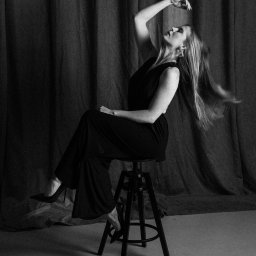STUDIO KREATYWNE Katarzyna Ćwiertnia - Czarno-białe zdjęcie kobiety w kombinezonie siedzącej na stołku barowym, z rozwianymi włosami i uniesioną ręką na tle drapowanej tkaniny