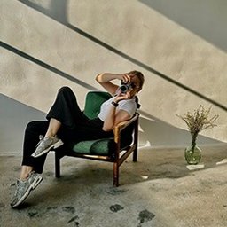 STUDIO KREATYWNE Katarzyna Ćwiertnia - Kobieta w okularach przeciwsłonecznych siedzi w zielonym fotelu, trzymając aparat i robiąc zdjęcie w jasnym pomieszczeniu z betonową podłogą i wazonem z suchymi kwiatami w tle.