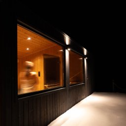 STUDIO KREATYWNE Katarzyna Ćwiertnia - Nowoczesna, drewniana sauna z dużymi oknami, oświetlona z zewnątrz, z widoczną osobą w środku i śniegiem na zewnątrz.