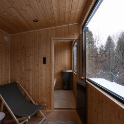 STUDIO KREATYWNE Katarzyna Ćwiertnia - Drewniana sauna z leżakiem i widokiem na zasypany śniegiem las przez duże okno, piec do sauny widoczny w głębi.