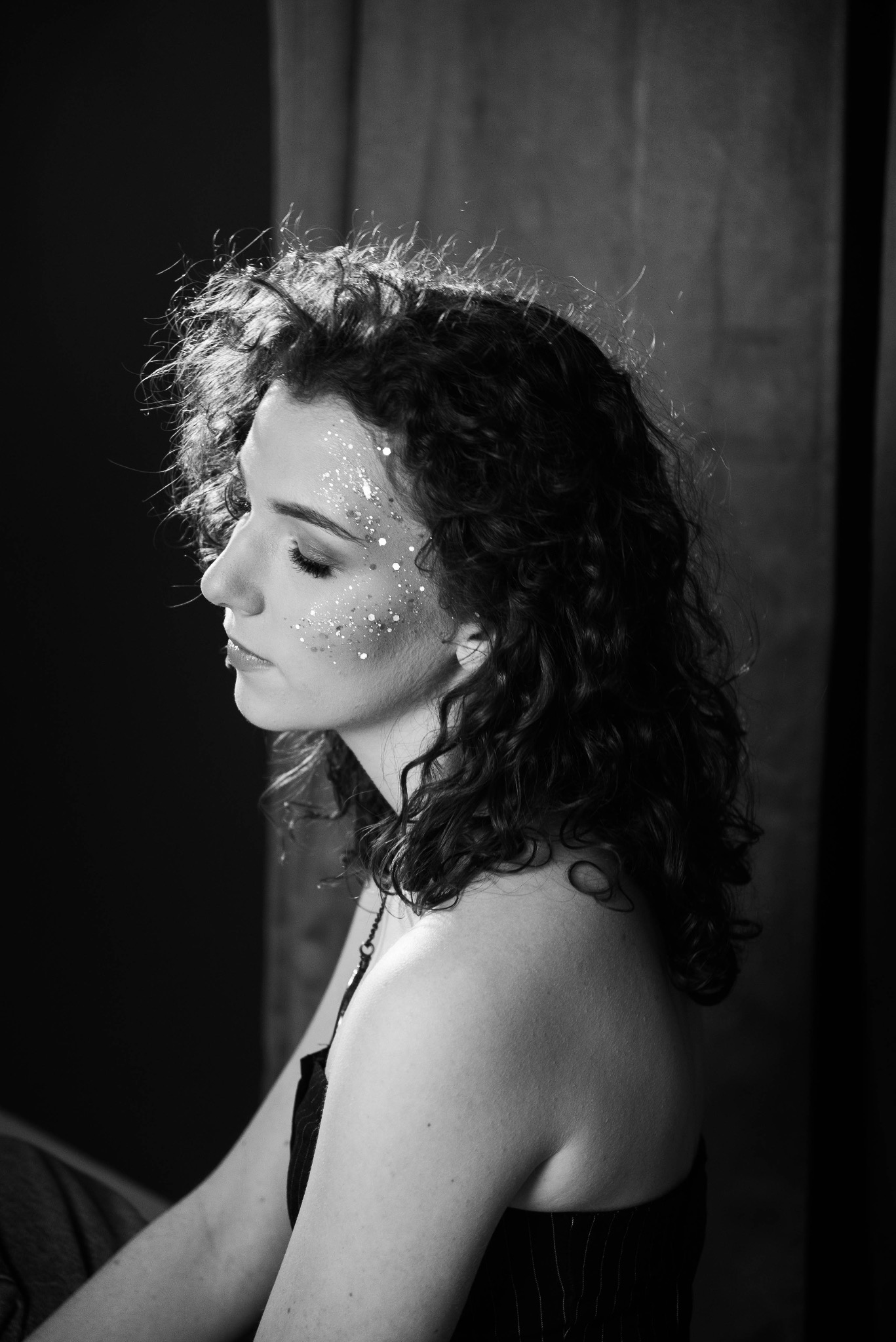 Czarno-białe portretowe ujęcie profilu kobiety z kręconymi włosami i brokatem na twarzy, w zamyślonej pozie na tle ciemnych zasłon.