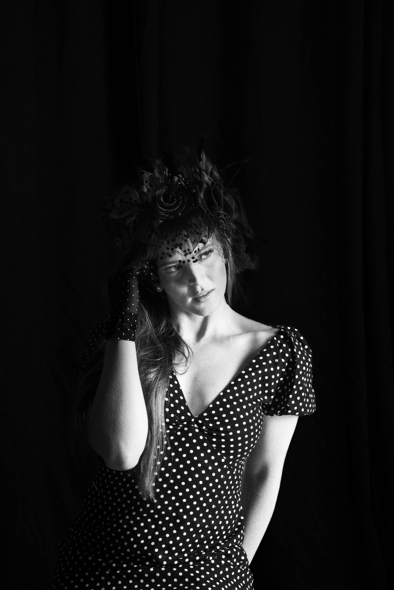 Czarno-białe portretowe ujęcie kobiety w sukience w grochy, z rękawiczkami w grochy i kapeluszem z woalką, pozującej na ciemnym tle.