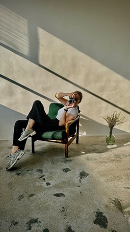 Kobieta w okularach przeciwsłonecznych siedzi w zielonym fotelu, trzymając aparat i robiąc zdjęcie w jasnym pomieszczeniu z betonową podłogą i wazonem z suchymi kwiatami w tle.