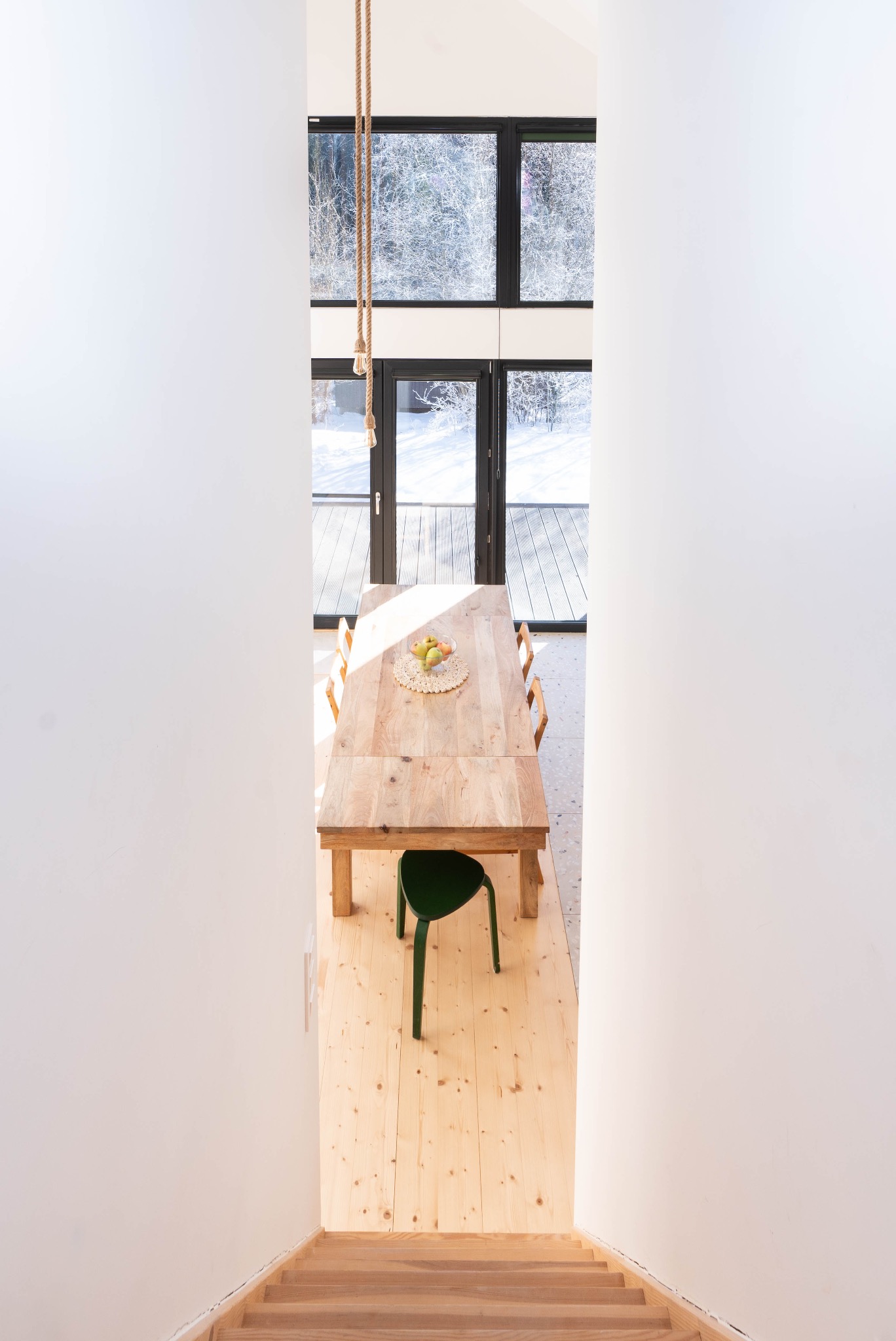 Wnętrze domu z drewnianym stołem, zielonym krzesłem i widokiem na zaśnieżony krajobraz przez duże okno, widziane z góry ze schodów.