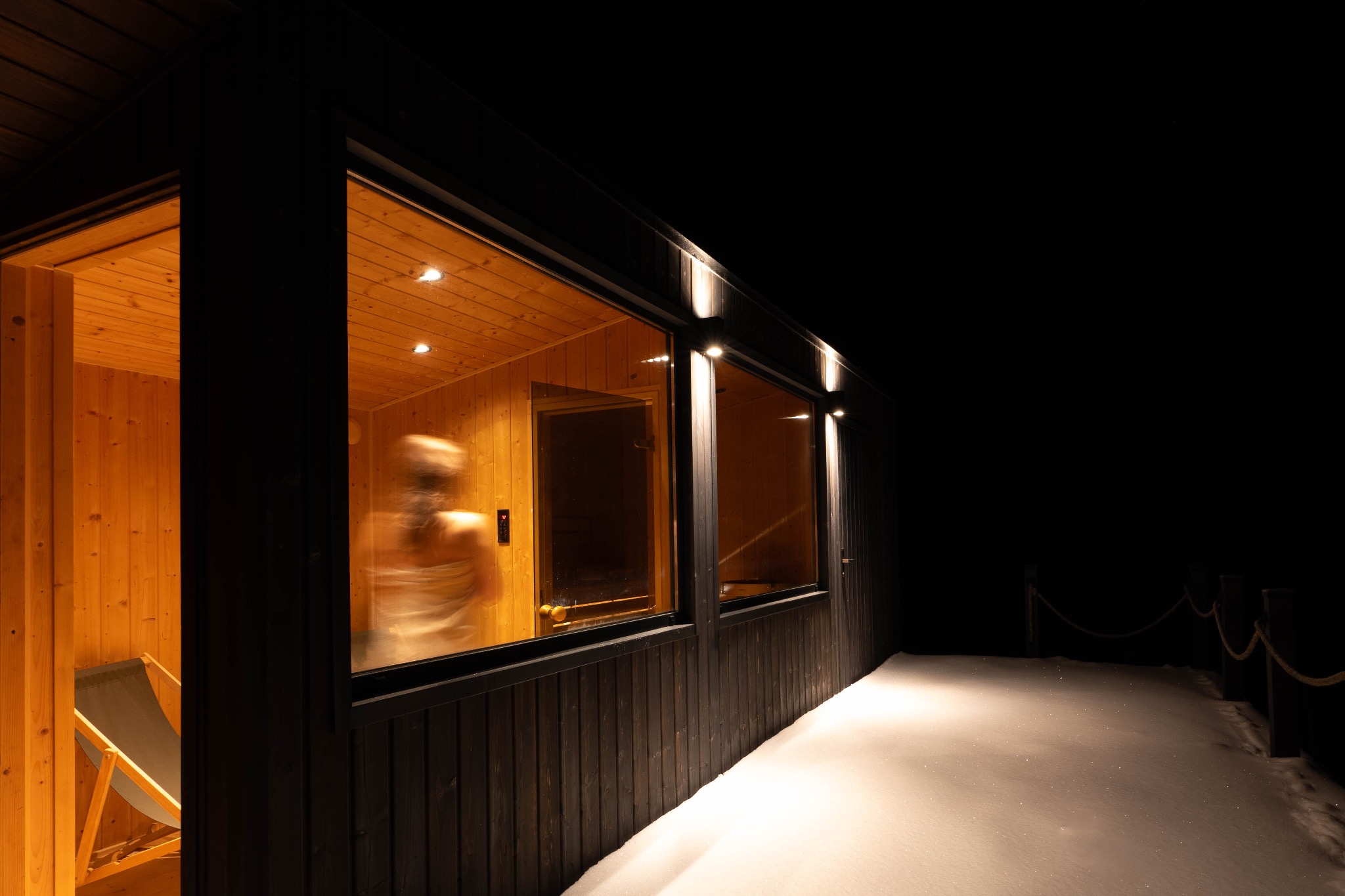 Nowoczesna, drewniana sauna z dużymi oknami, oświetlona z zewnątrz, z widoczną osobą w środku i śniegiem na zewnątrz.
