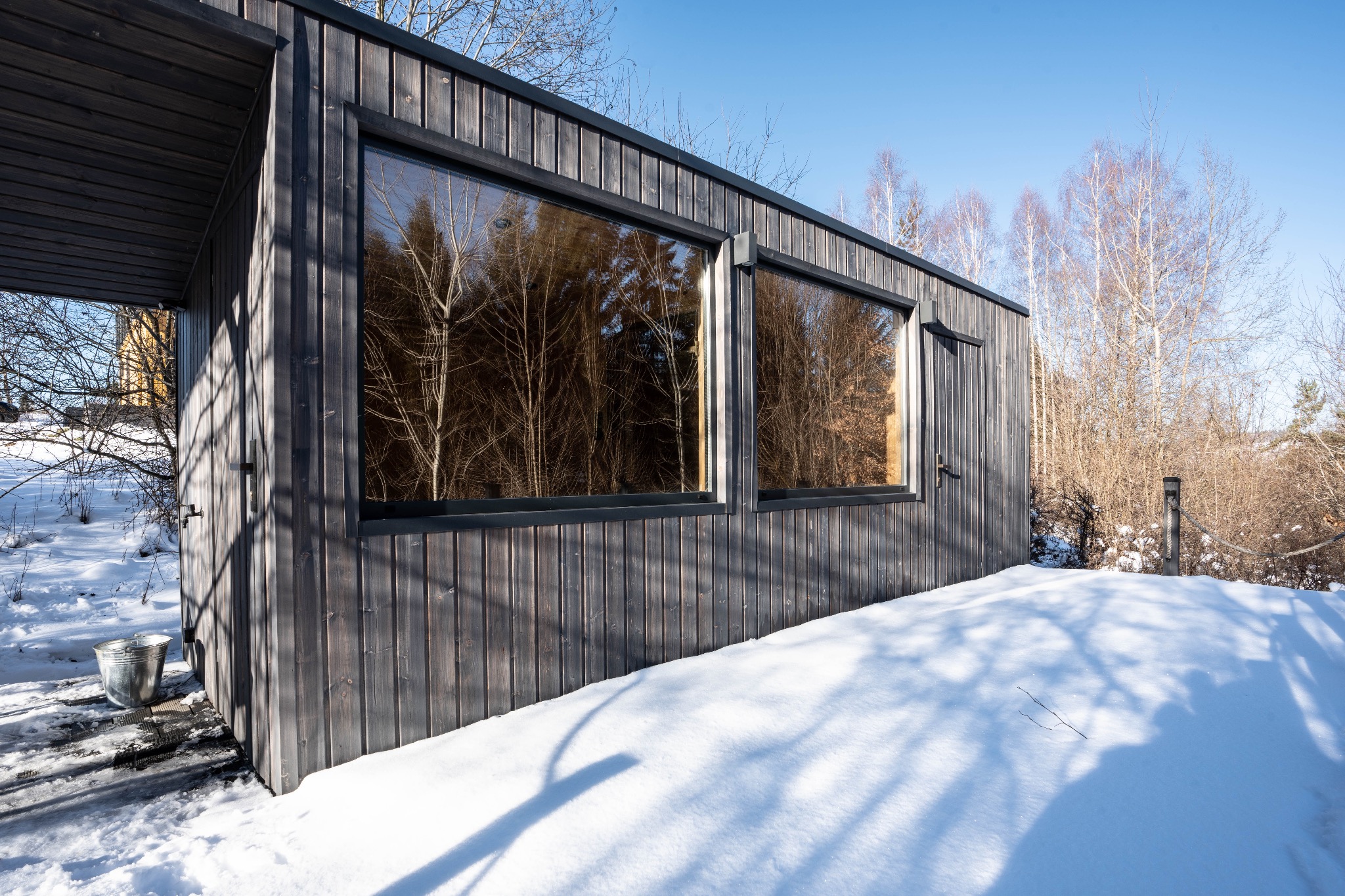 Nowoczesny, ciemny, drewniany domek w zimowej scenerii, z dwoma dużymi oknami odbijającymi drzewa i błękitne niebo, z metalowym wiadrem stojącym obok wejścia, otoczony śniegiem.