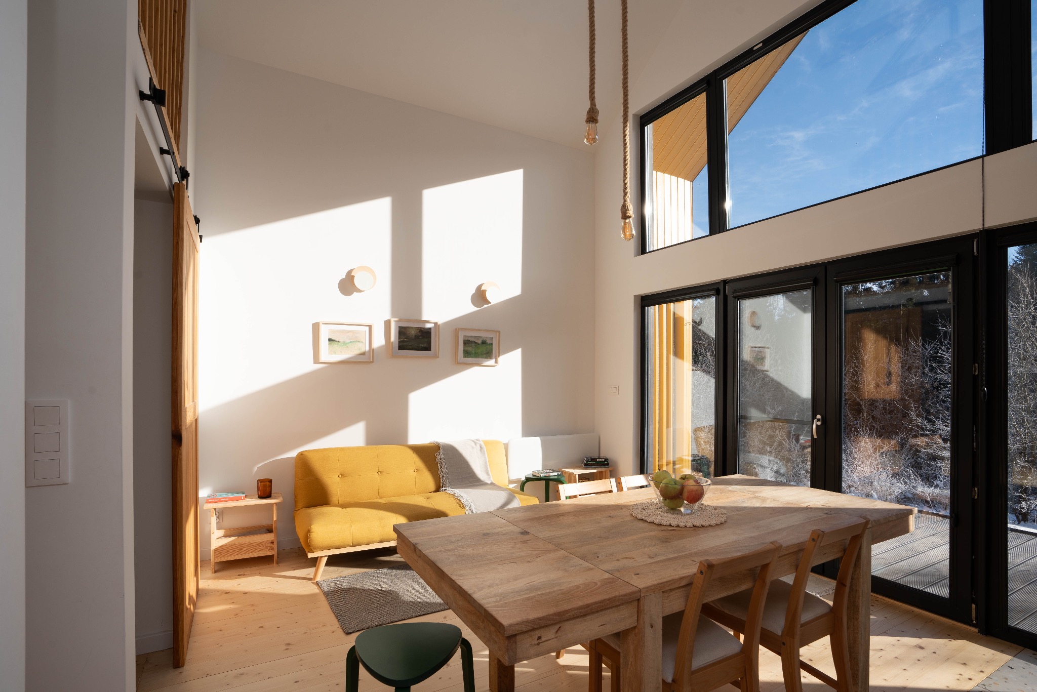 Jasne wnętrze domu z drewnianą podłogą, żółtą sofą, drewnianym stołem z owocami i dużymi oknami z widokiem na zewnątrz, oświetlone naturalnym światłem.