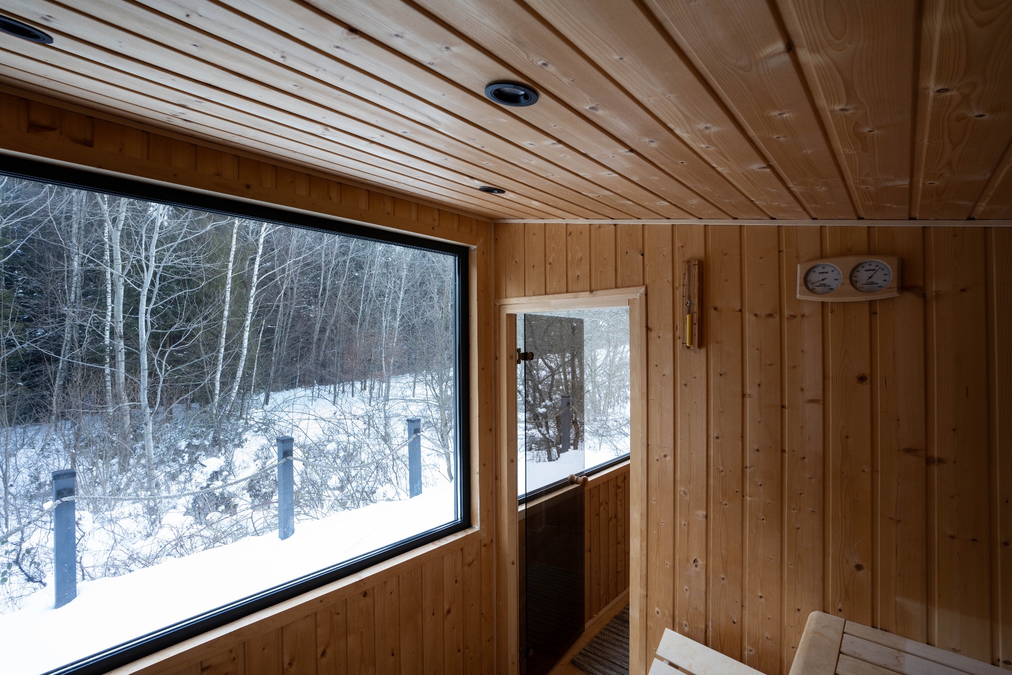 Wnętrze sauny z drewnianymi ścianami, sufitem i ławkami, duże okno z widokiem na zimowy krajobraz z drzewami i śniegiem, termometr i higrometr na ścianie.