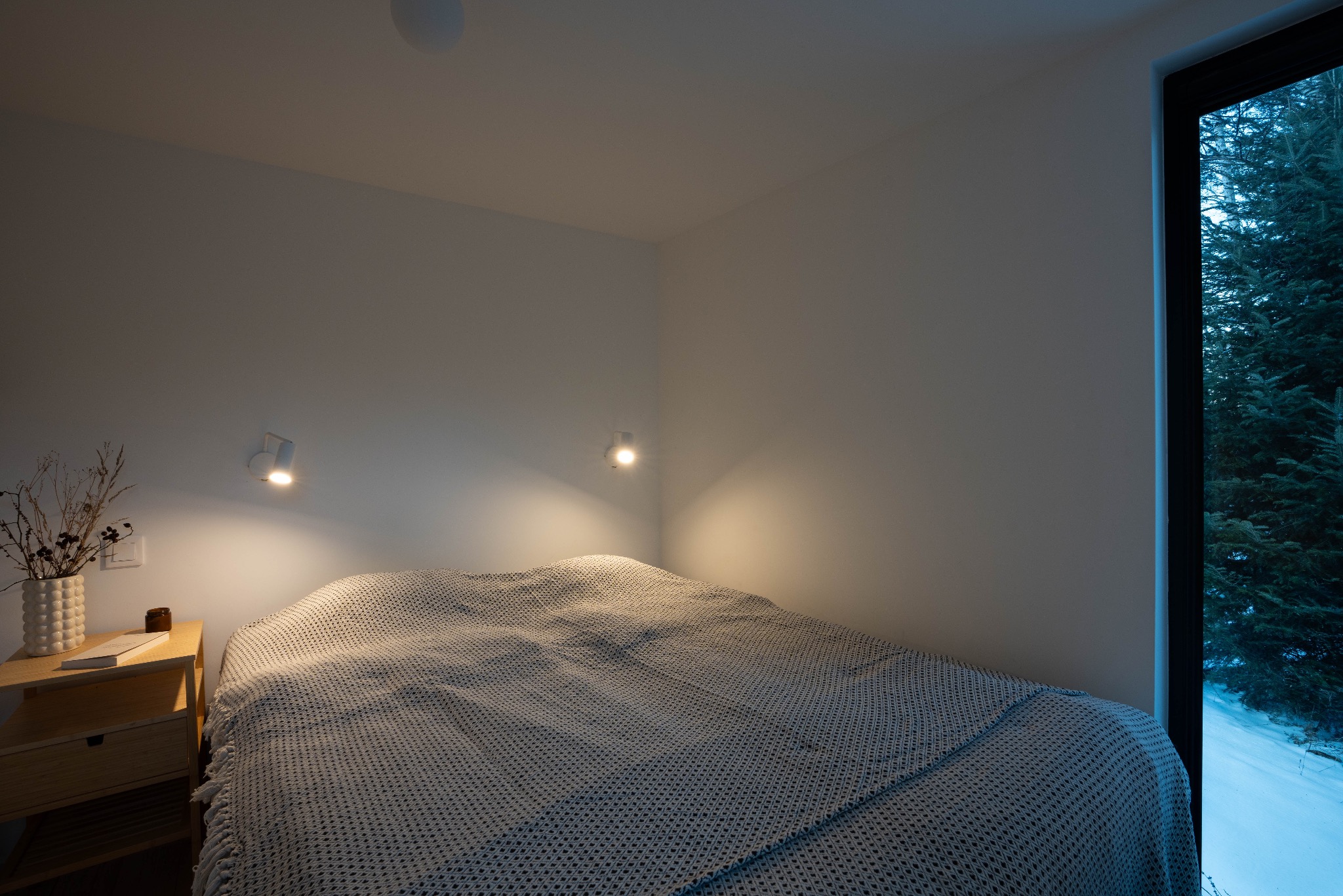 Jasna sypialnia z łóżkiem przy oknie z widokiem na zimowy las, na stoliku nocnym wazon z suchymi kwiatami i książka, oświetlenie punktowe.