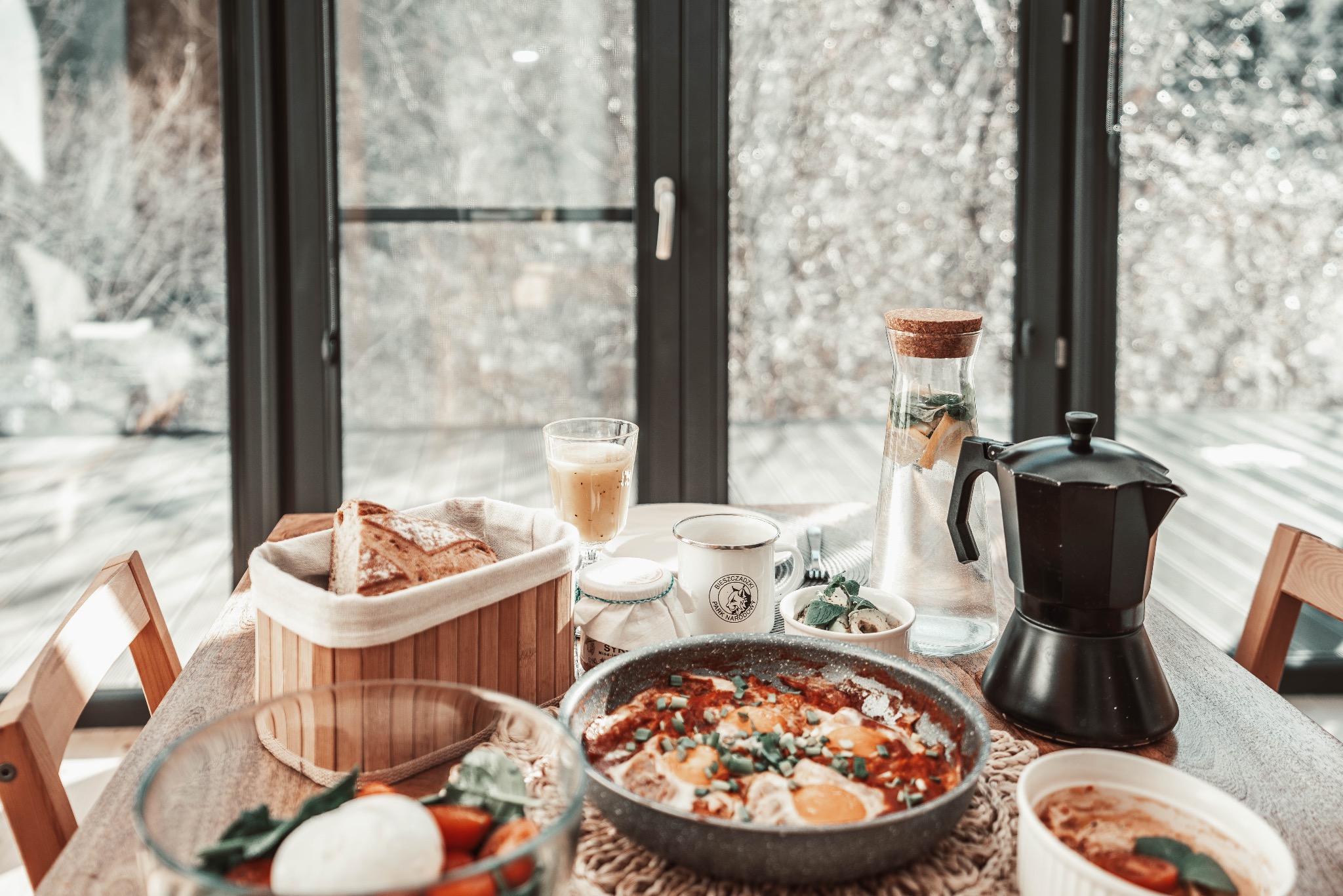 Śniadanie w Bieszczadach: Szakszuka w patelni, pieczywo w koszyku, kawa w kawiarce, lemoniada w karafce i sok na drewnianym stole w @rownownoc_w_bieszczadach.